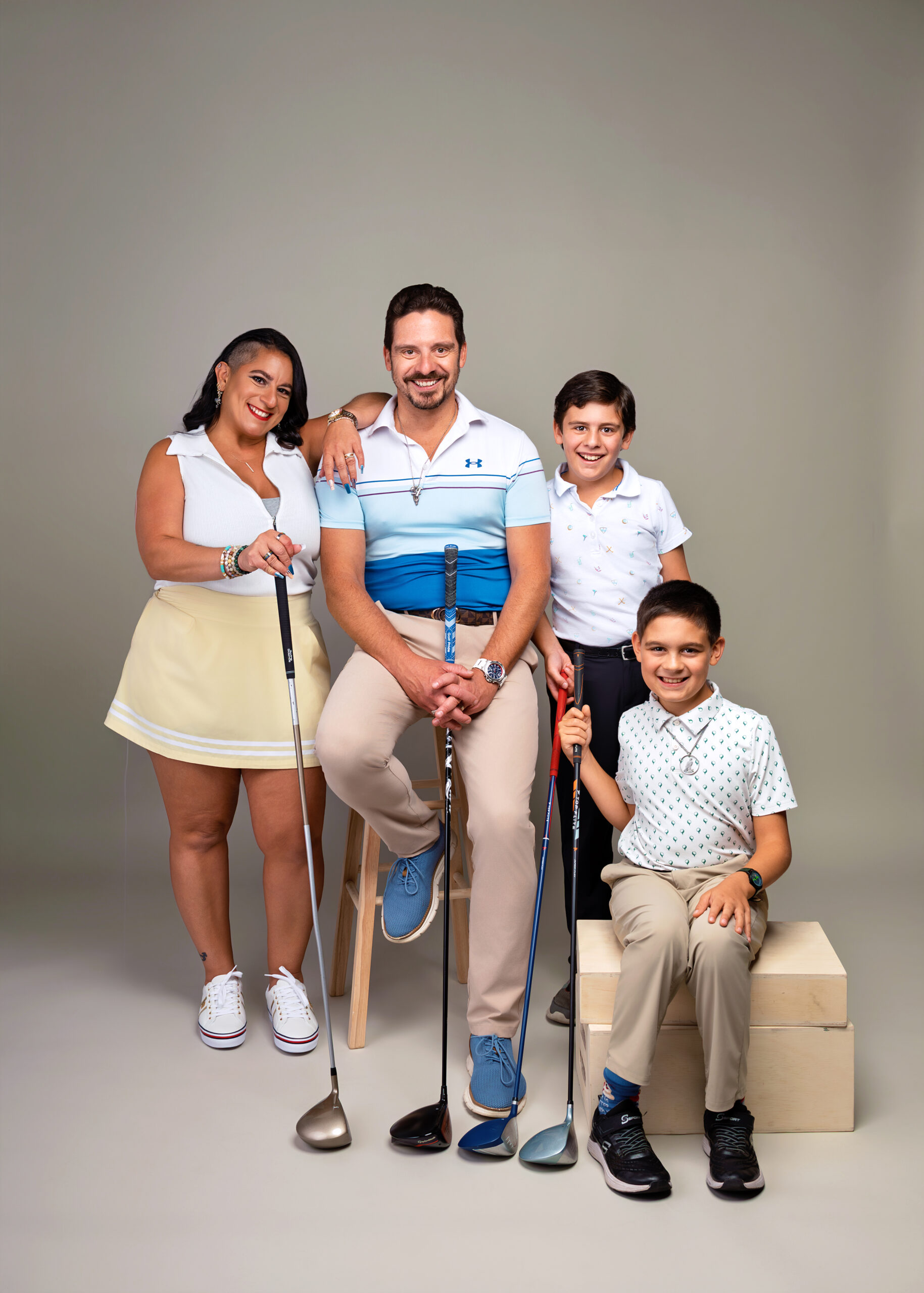 Family of four posing with golf clubs in a studio; mother on the left, father center, two boys on the right, all smiling at the camera.