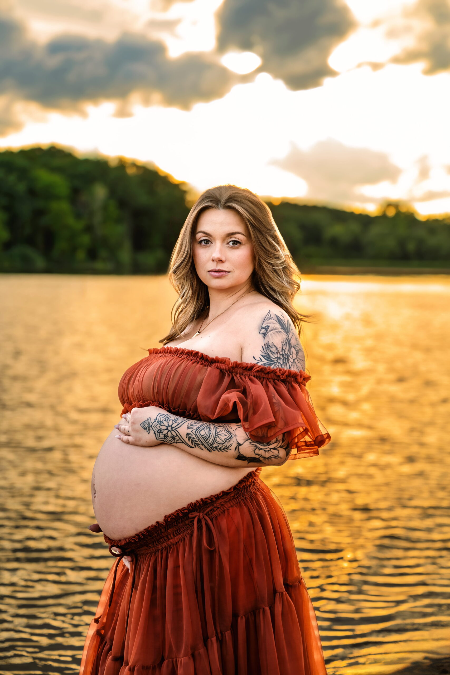 Pregnant woman in an orange off-shoulder dress stands by a lake at sunset, with visible arm tattoos.