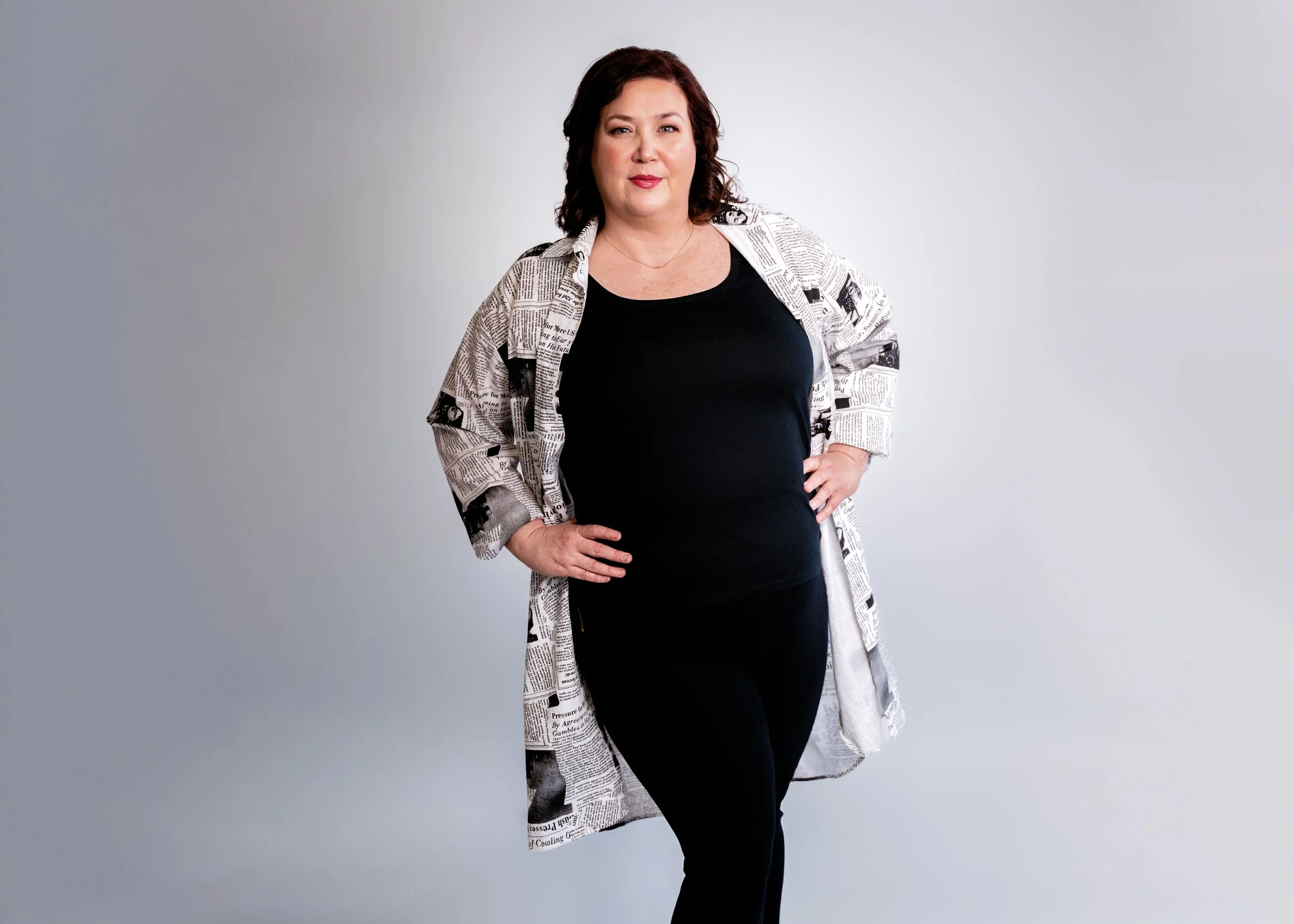 A woman with shoulder-length dark hair, wearing a black top and pants, and a newspaper print coat, standing with hands on hips against a plain light gray background.