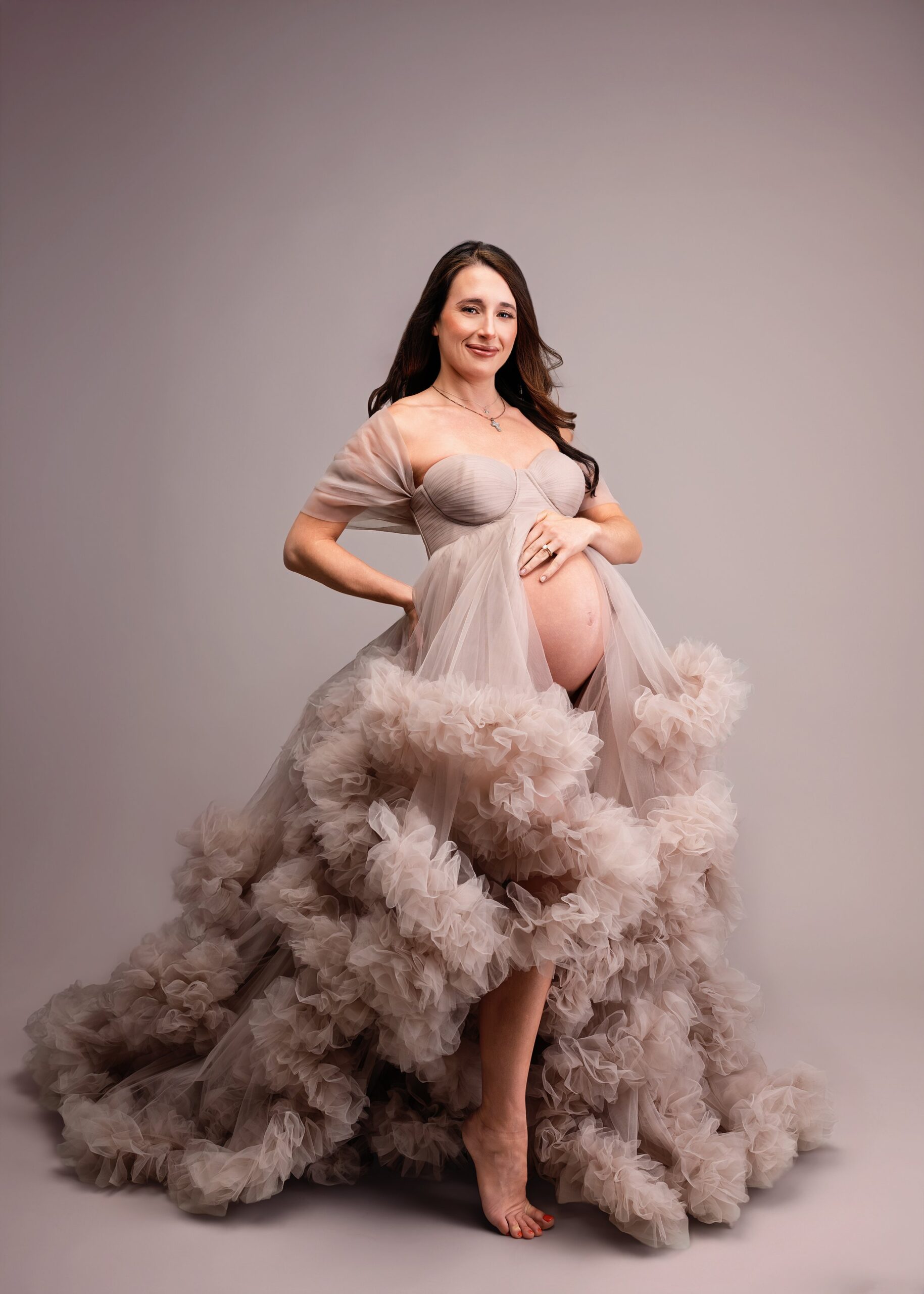 A pregnant woman standing against a plain background, wearing a nuptial-style, ruffled, beige dress and smiling at the camera.