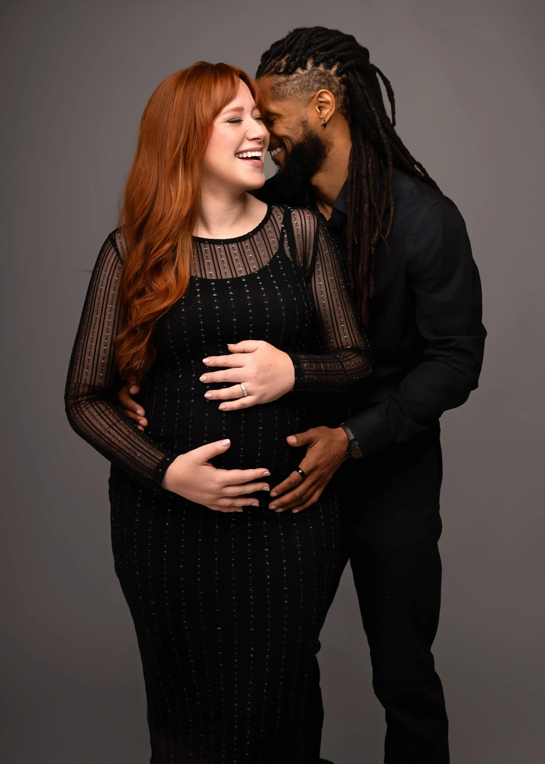 A joyful couple, a pregnant woman with red hair and a man with dreadlocks, affectionate together, with their foreheads touching and hands on her belly, against a plain gray background.
