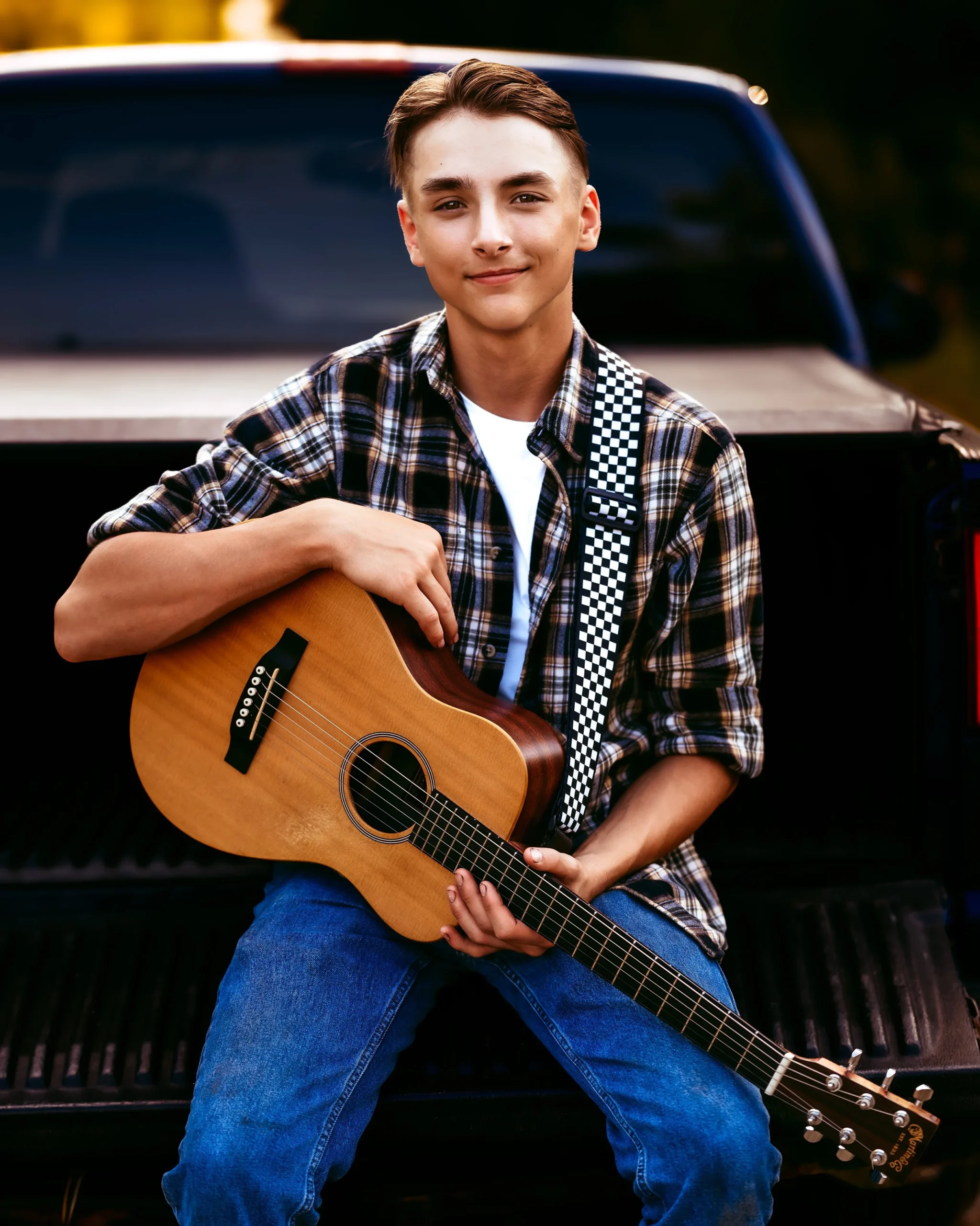 Young man with guitar sitting on a truck bed, wearing a plaid shirt and jeans.