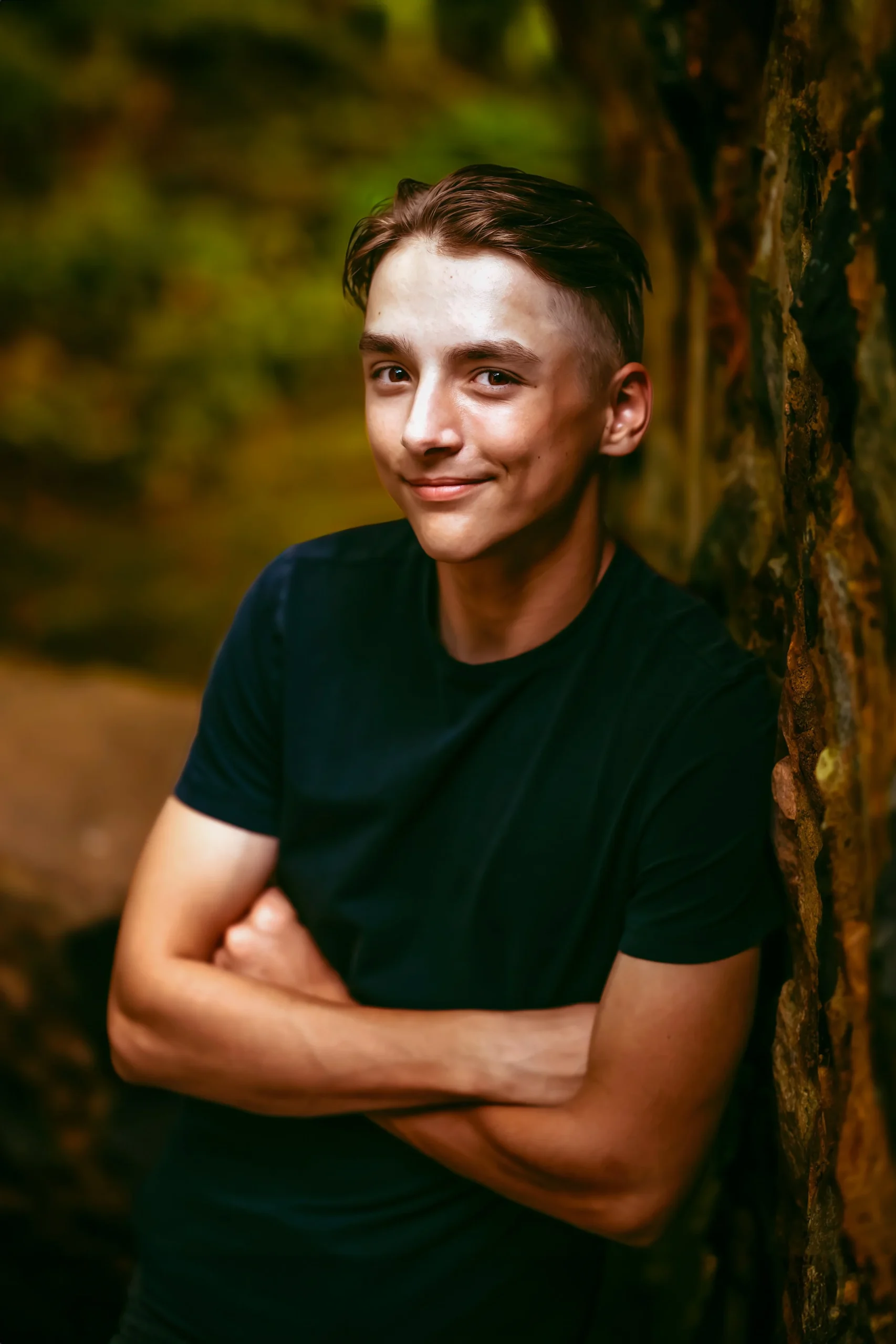 Young man with a confident smile, wearing a black T-shirt, standing with arms crossed against a textured wall, with a blurred natural background.