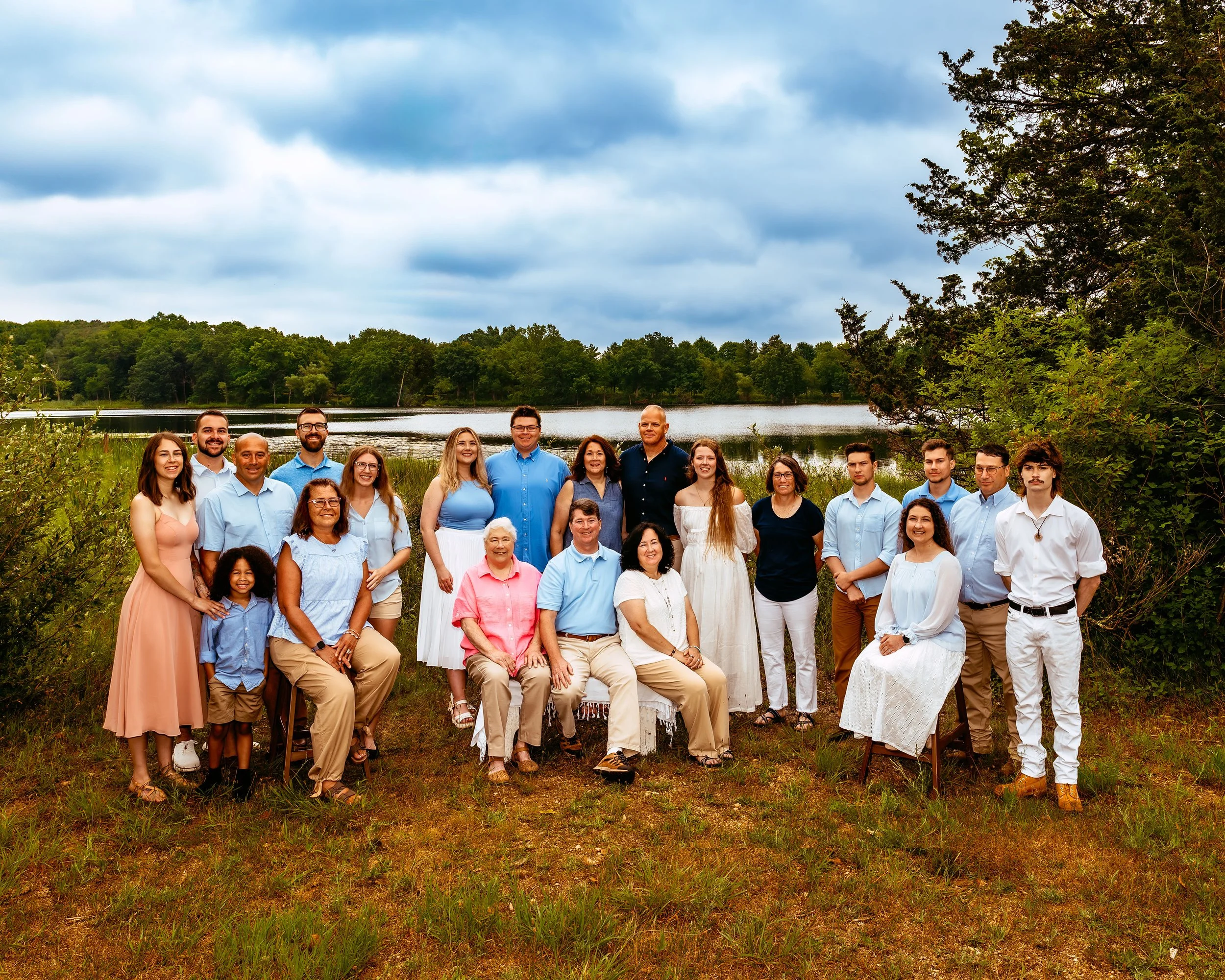 A group of people posing for a family portrait outdoors near a lake, with greenery and a cloudy sky in the background. They are dressed in casual clothing, with color-coordinated outfits consisting mostly of blue, white, and pastel tones. The group i