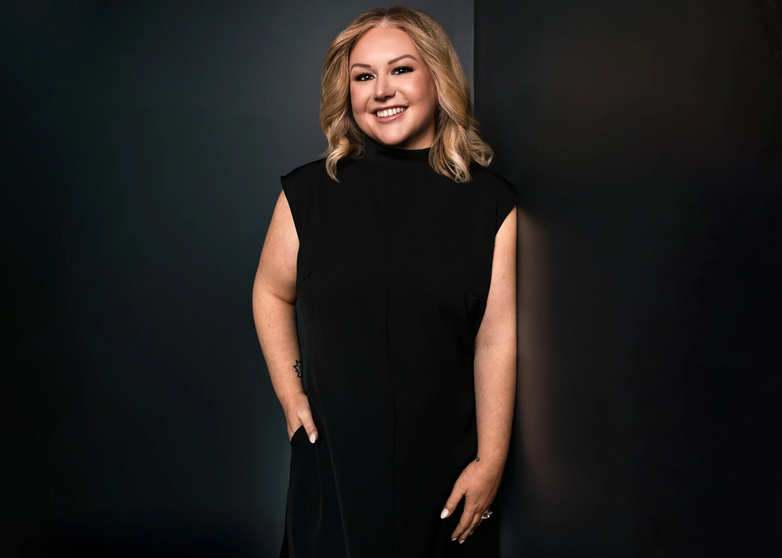 A smiling person with blonde hair wearing a sleeveless black dress stands against a dark background with hands in pockets.