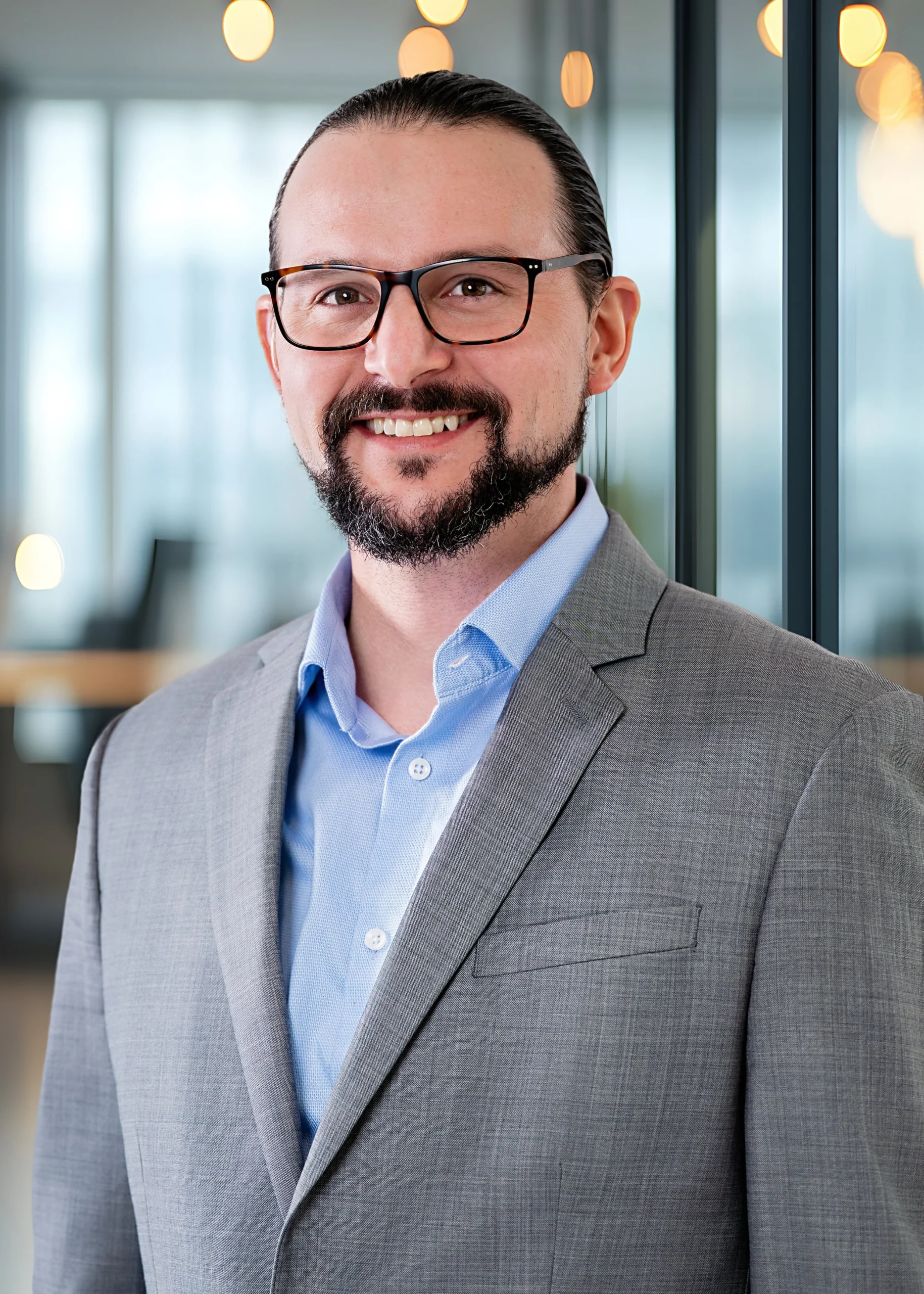 A man with glasses and a beard, wearing a gray suit and blue shirt, smiling in an office setting.