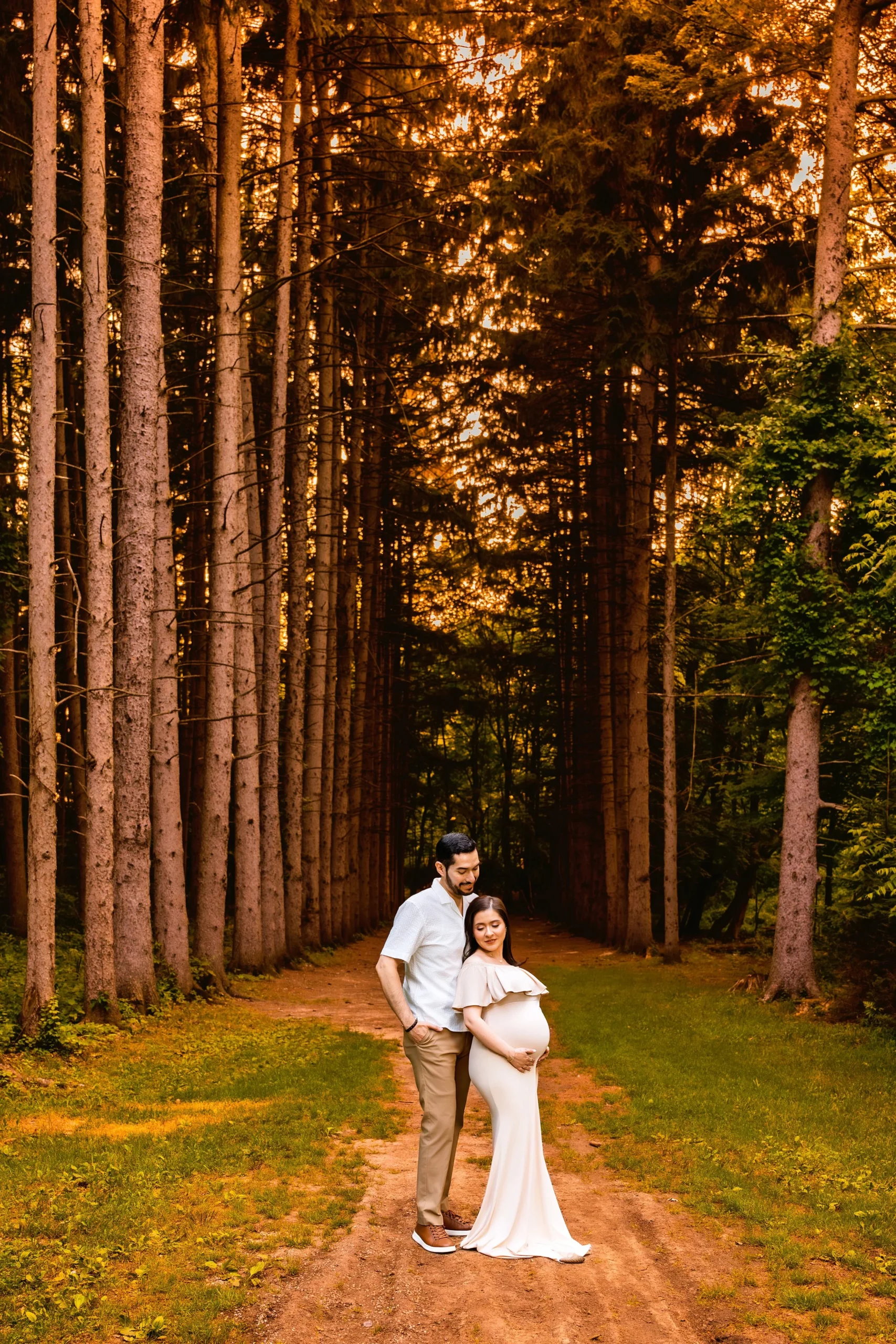 A pregnant woman in a white dress standing on a forest path with a man, surrounded by tall trees, during sunset.