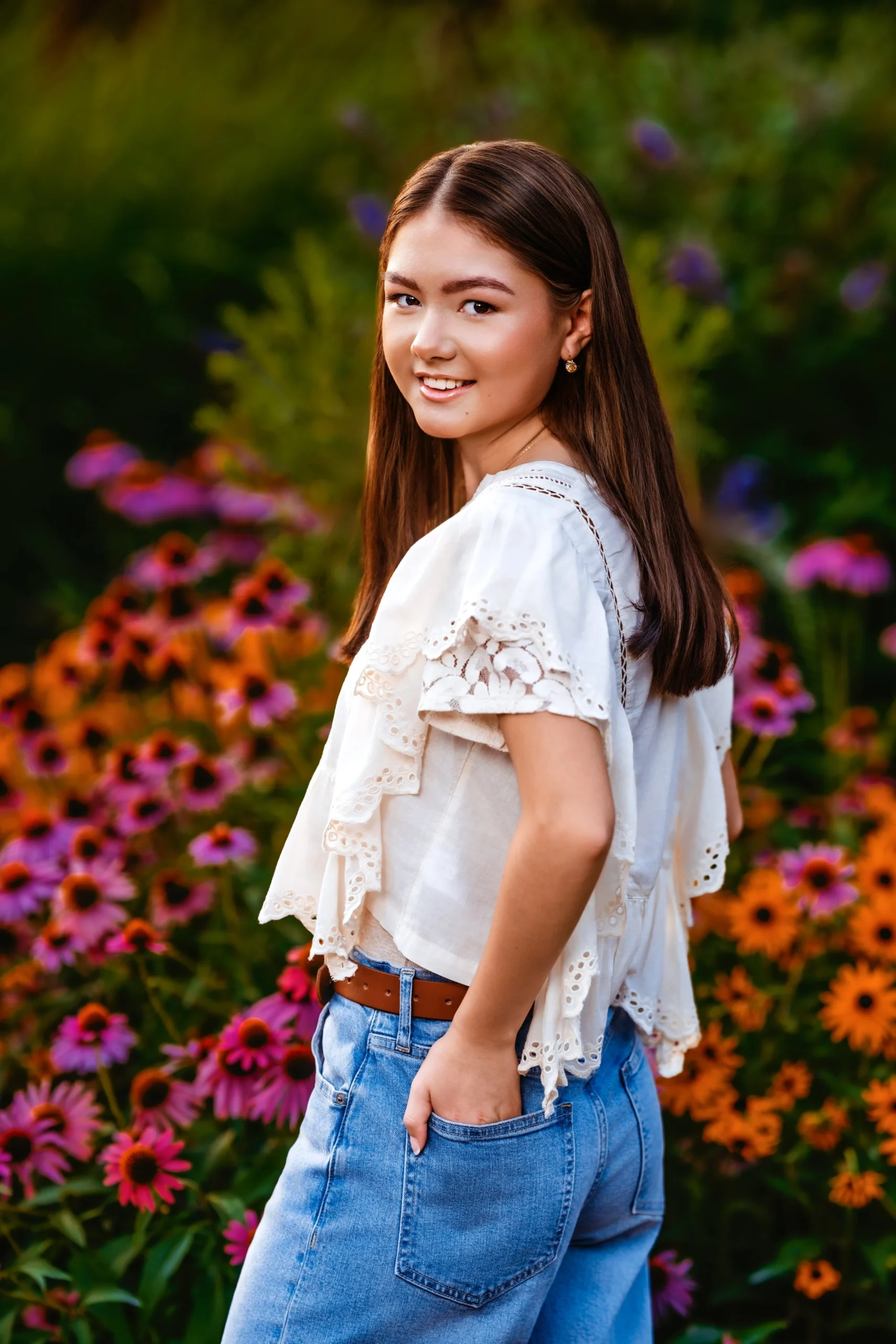 Young woman with long brown hair smiling in a garden filled with pink and orange flowers.