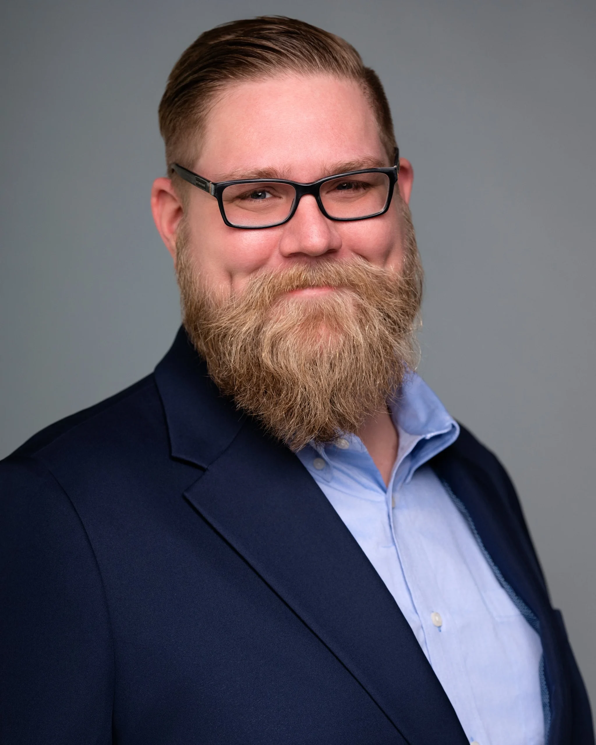 A man with glasses, a beard, and a mustache, wearing a dark blazer over a light blue shirt, smiling at the camera.