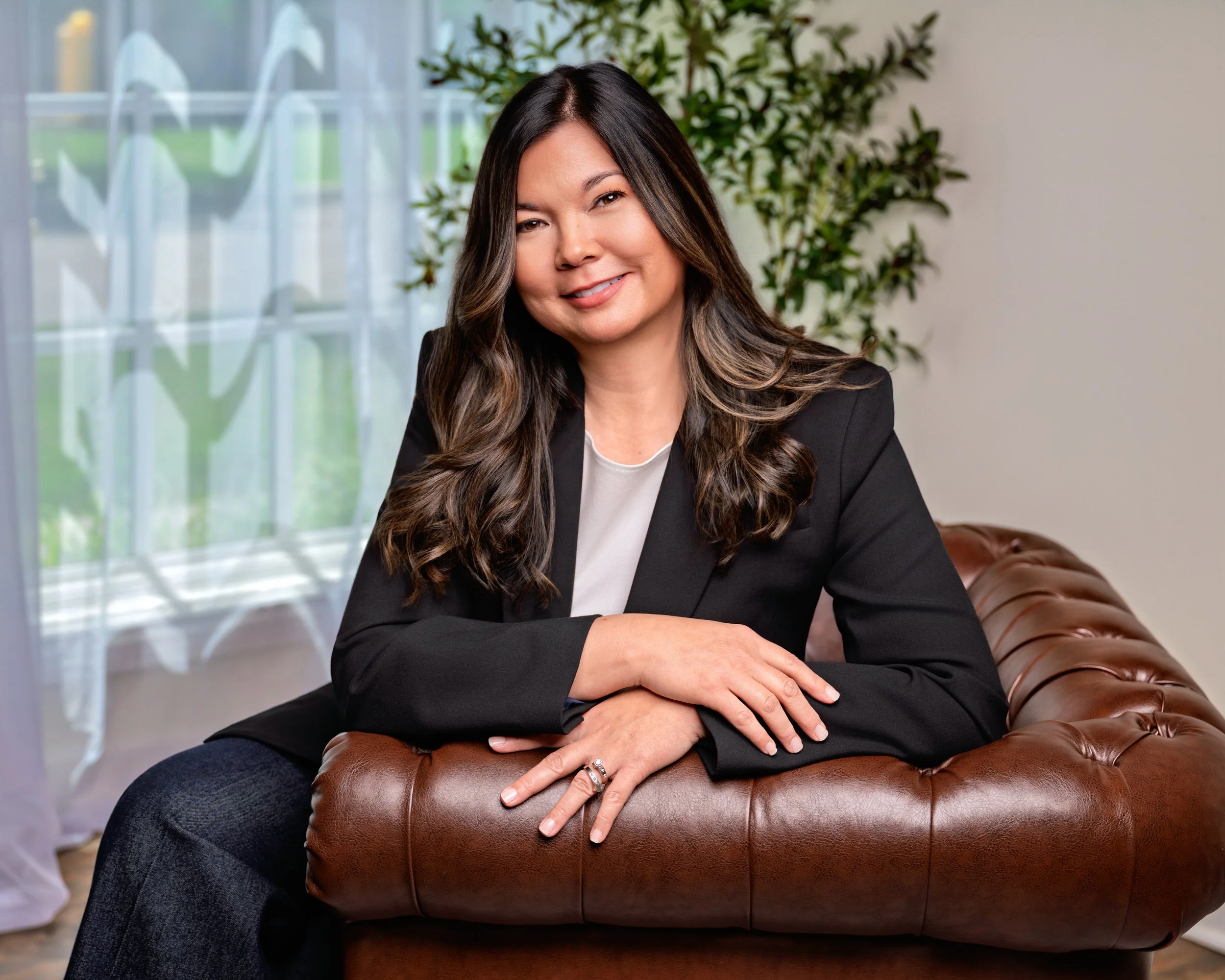 A woman with long dark hair and a black blazer sitting on a brown leather couch in a well-lit room with a window, sheer curtains, and a potted plant in the background.
