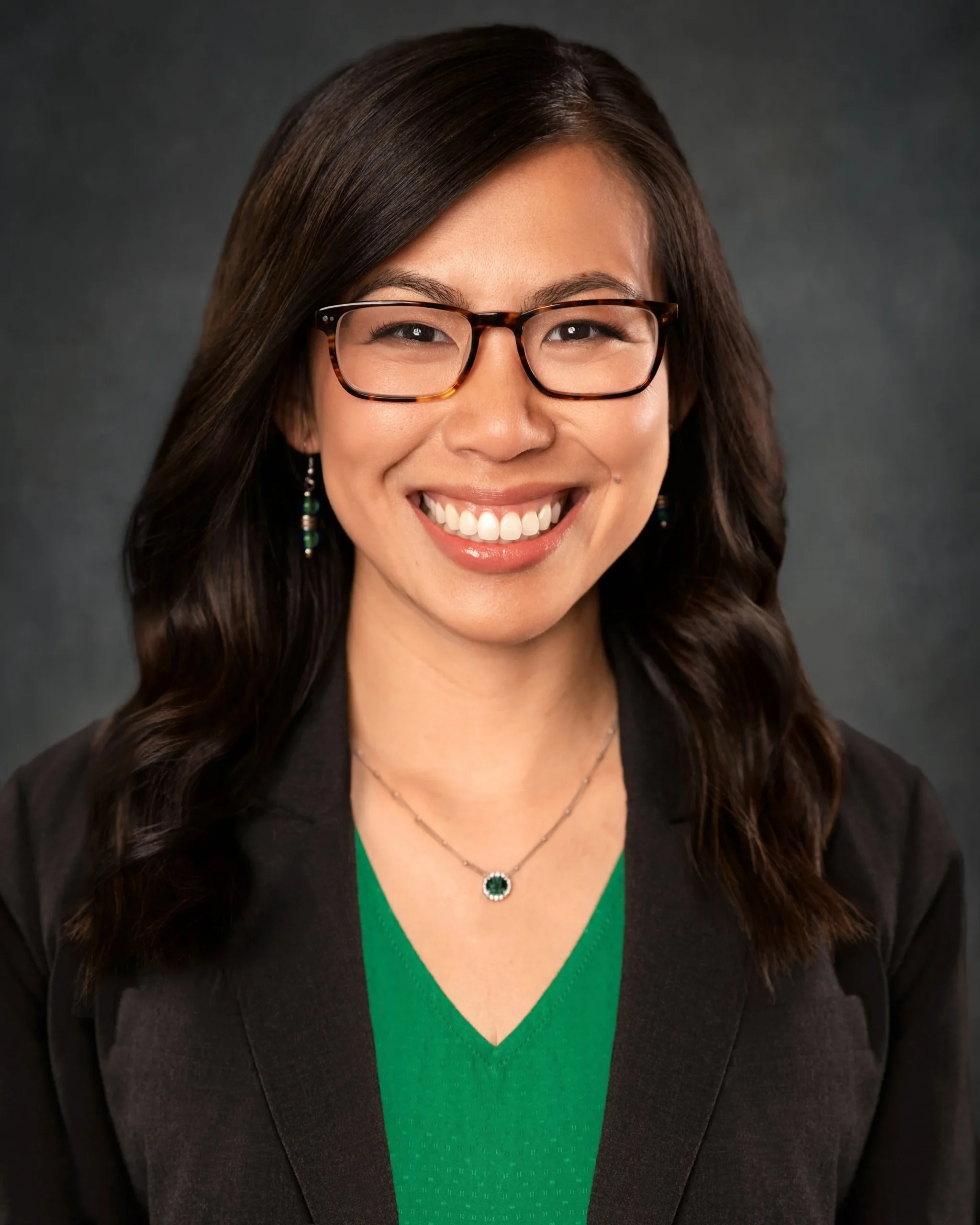 A woman with long dark hair, glasses, and a bright smile, wearing a green top, a black blazer, and jewelry, against a dark background.