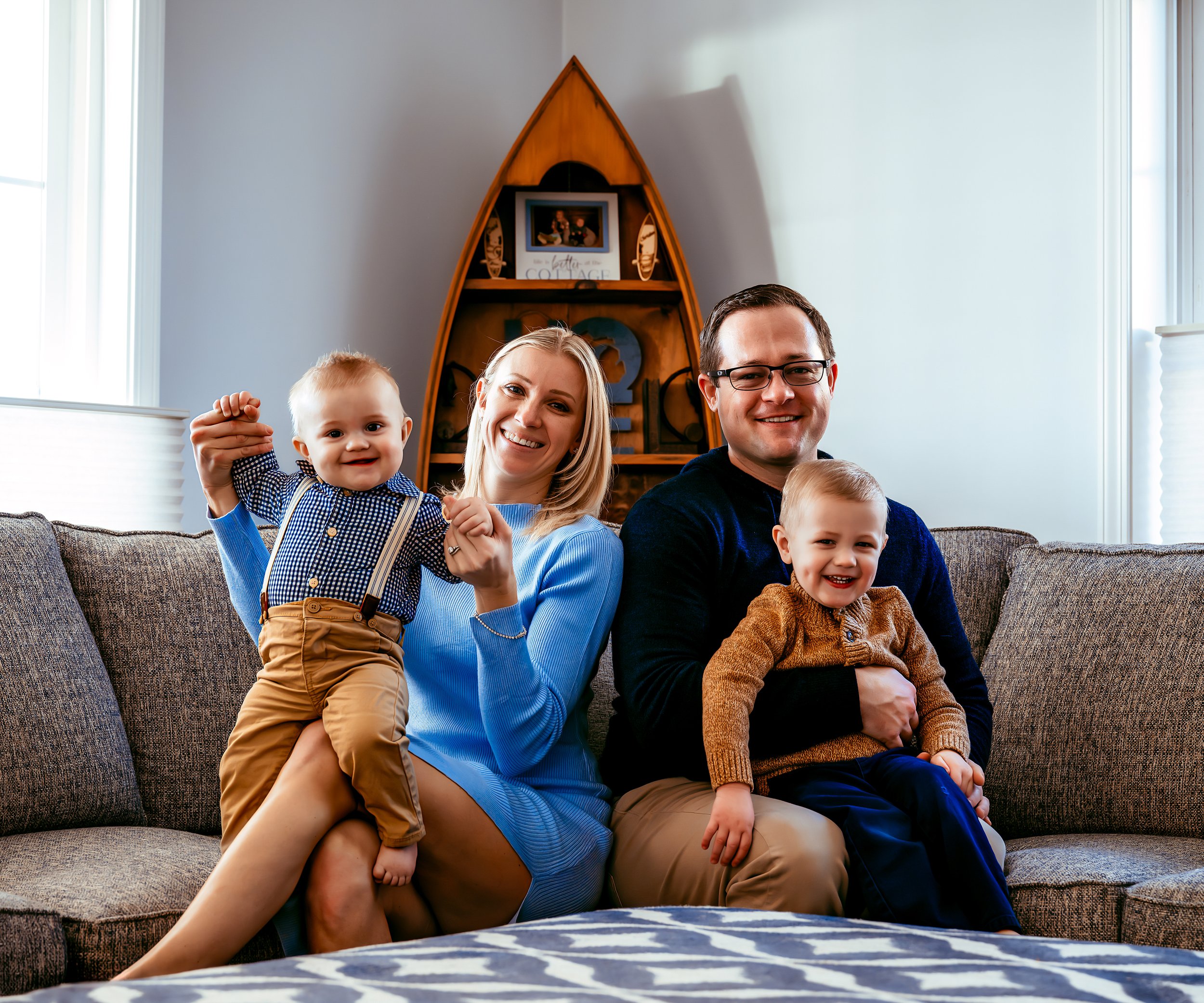 A family of four sitting on a couch, with parents holding two young children. There is a boat-shaped bookshelf in the background.
