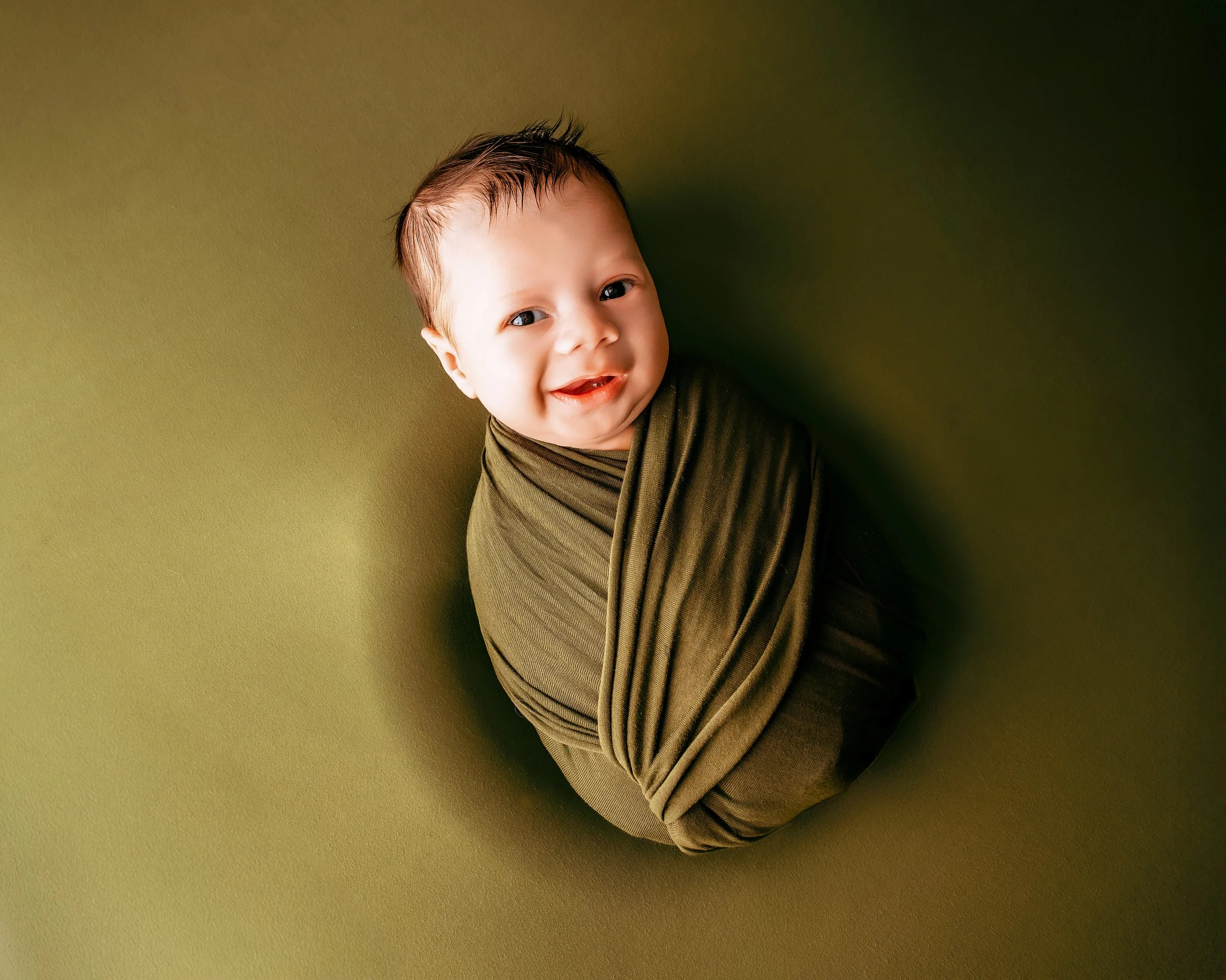 Smiling baby with short brown hair wrapped in a green cloth, lying on a green background.