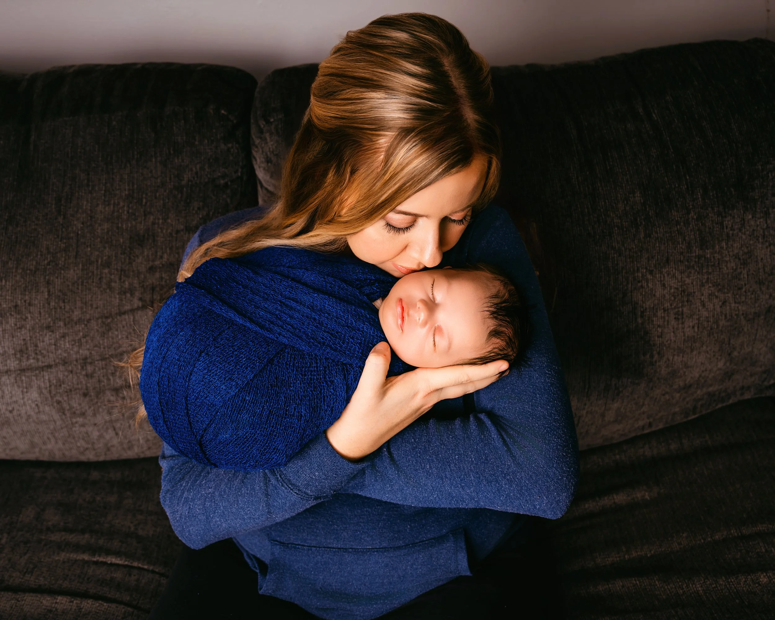 A woman with long light brown hair holding a sleeping baby wrapped in a blue blanket on her lap, sitting on a dark sofa.