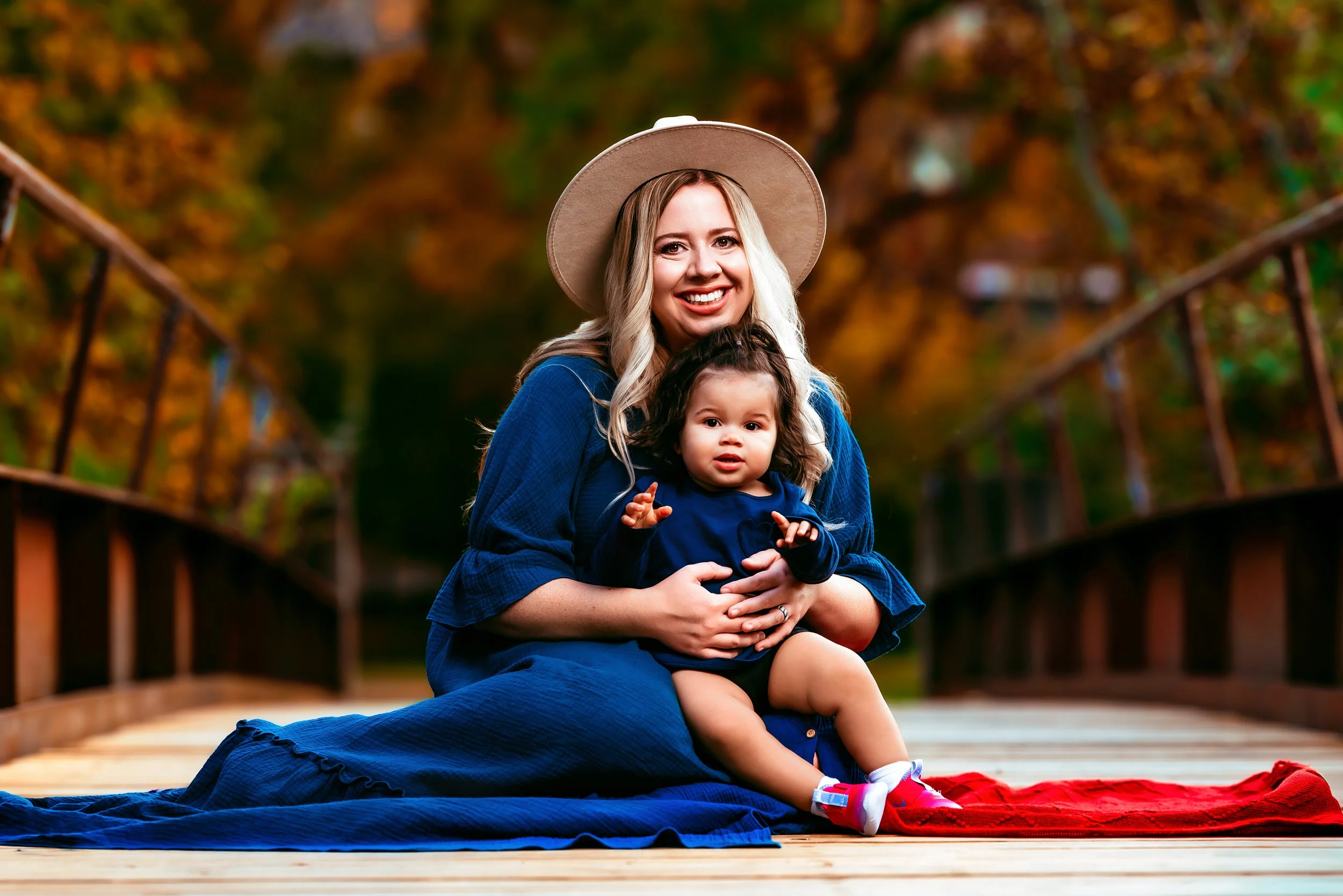 Woman in a blue dress and wide-brimmed hat holding a baby on a wooden bridge with fall foliage background.