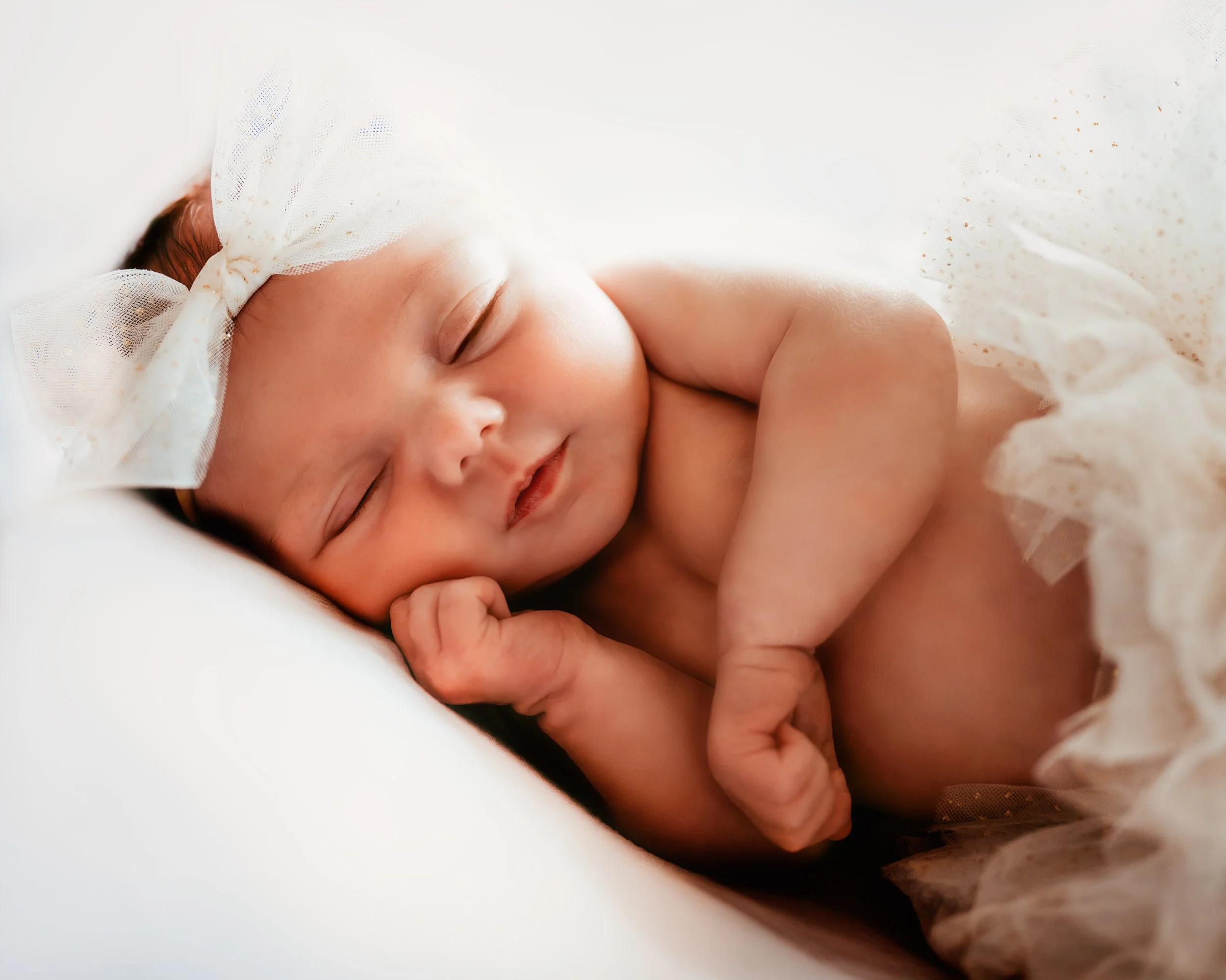 Sleeping newborn baby wearing a white bow headband and tulle skirt on a soft surface.