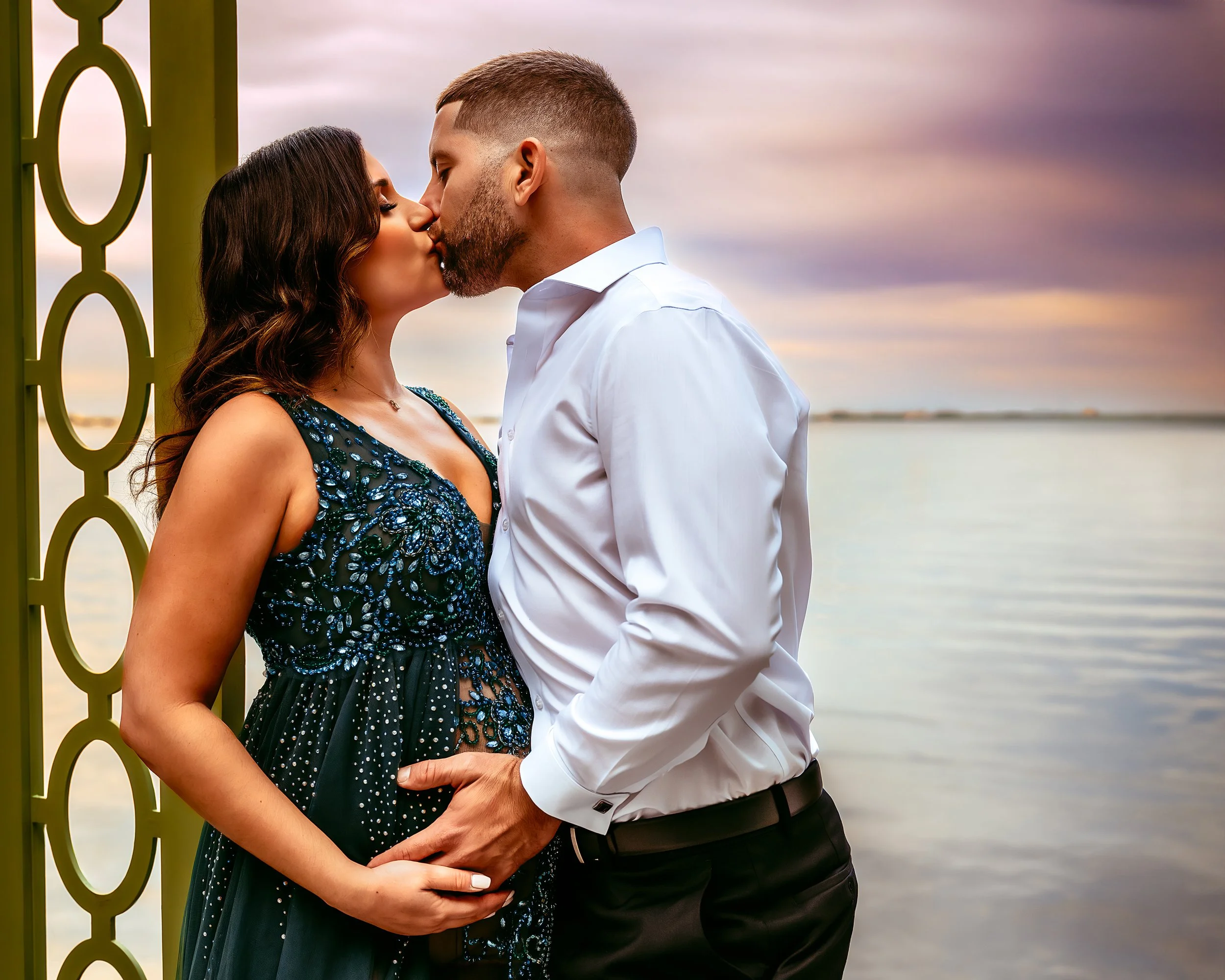 Couple kissing by the water during sunset, with the woman in a beaded dress and the man in a white shirt, their hands resting on her baby bump.