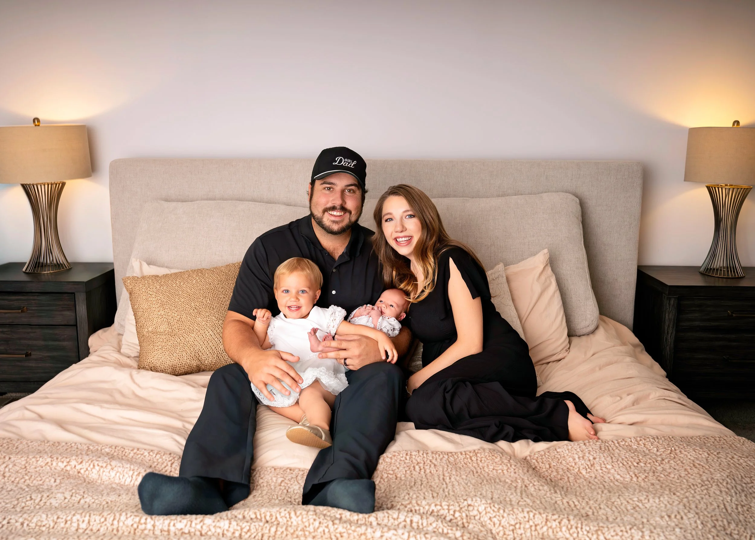 Family of four sitting on a bed, smiling at the camera, with two parents, a toddler girl, and a newborn baby girl, in a bedroom with bedside lamps and beige bedding.