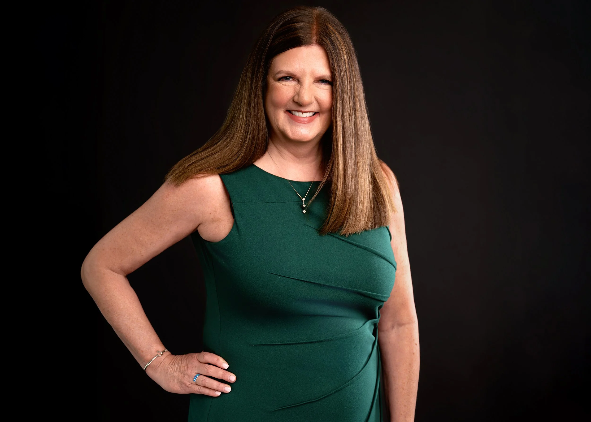 A smiling woman with long, straight brown hair wearing a sleeveless green dress, standing against a black background.