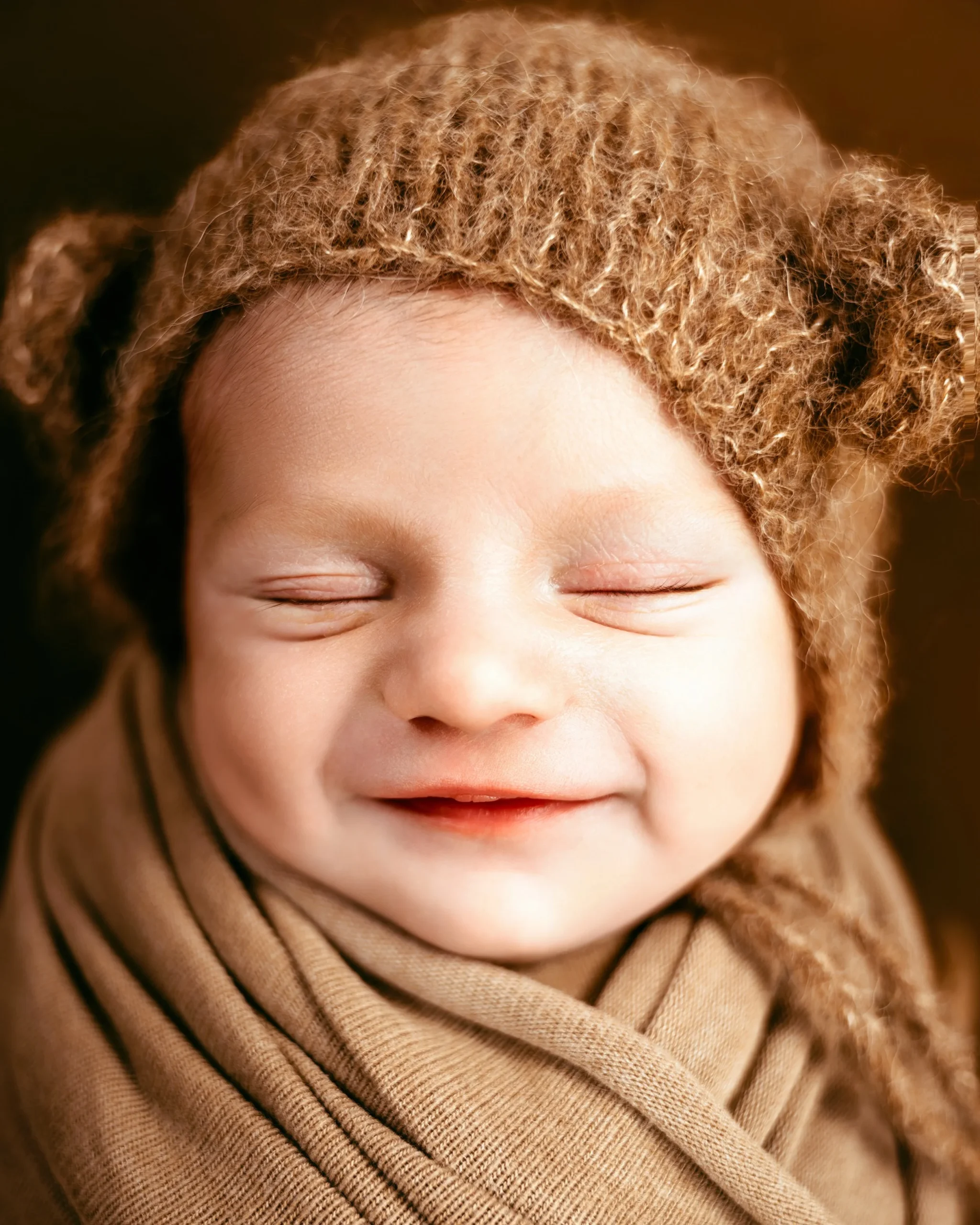 Close-up of a smiling baby with curly hair, closed eyes, and wearing a brown knit hat and scarf.