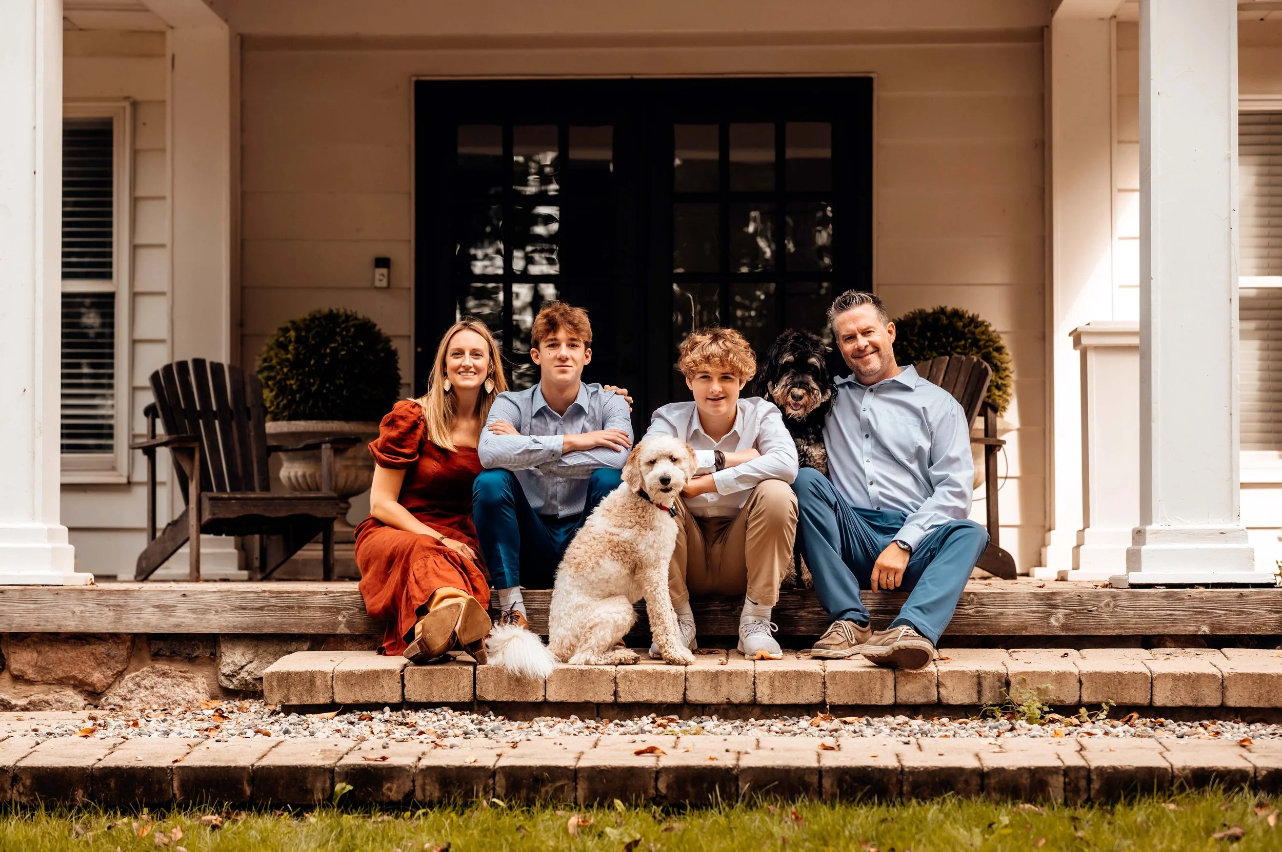 Family sitting on porch steps with two dogs, wearing casual clothes, in front of house with white siding and black door.