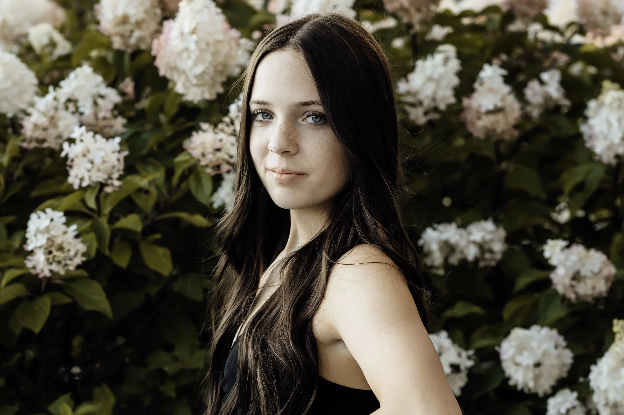 Young woman with long brown hair standing in front of white hydrangea flowers.