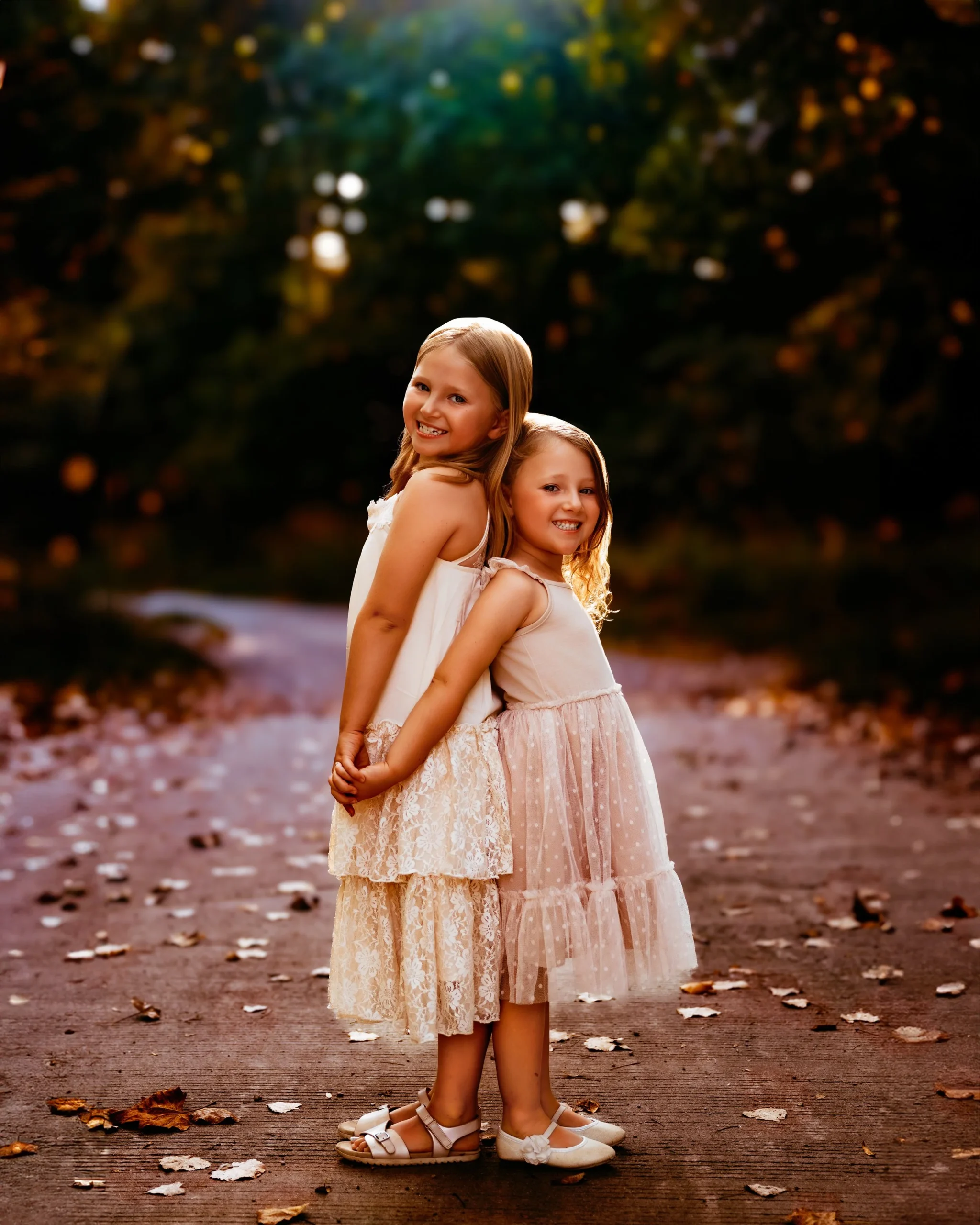 Two young girls in white dresses smiling, standing back-to-back on a path with autumn leaves.