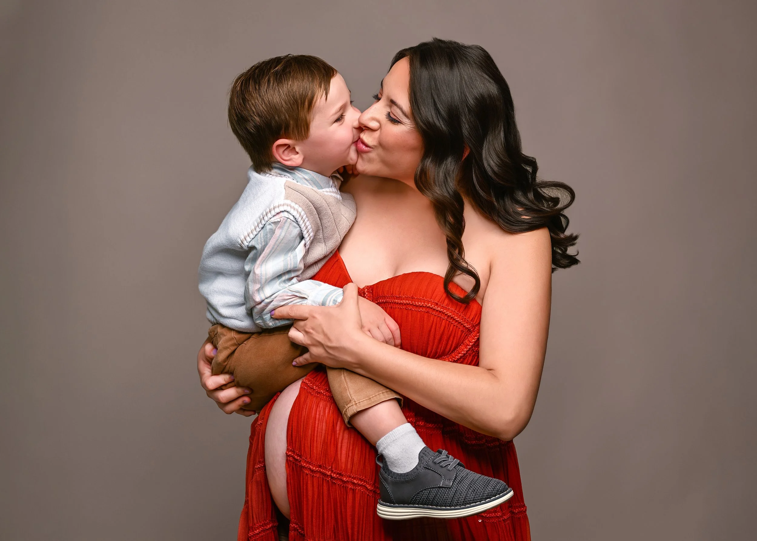 A woman with long dark hair and a red dress holding a young boy, giving him a kiss on the nose against a plain gray background.