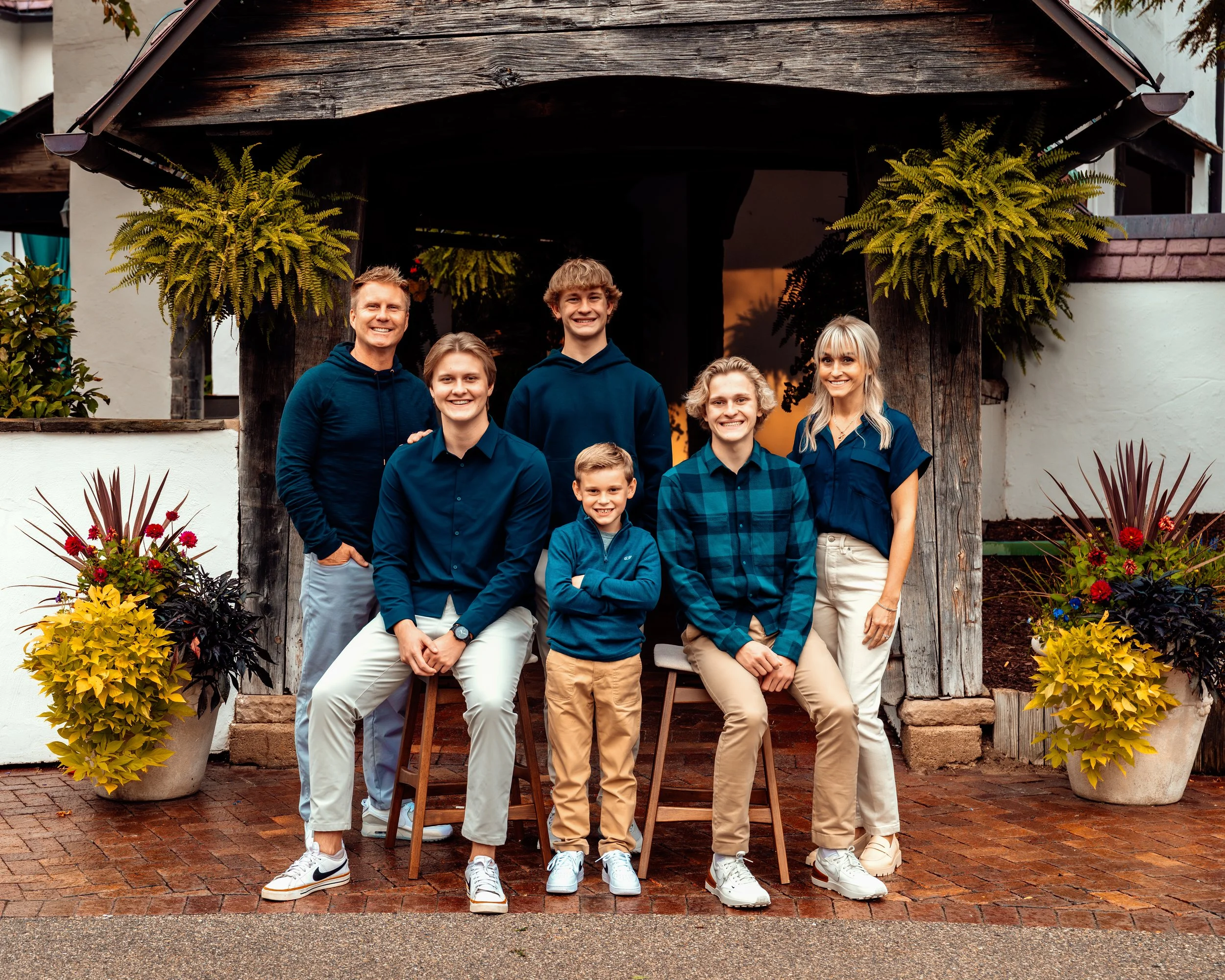 Family posing together in front of a rustic wooden structure, surrounded by hanging plants and floral arrangements. Adults and children are dressed in coordinated casual outfits.
