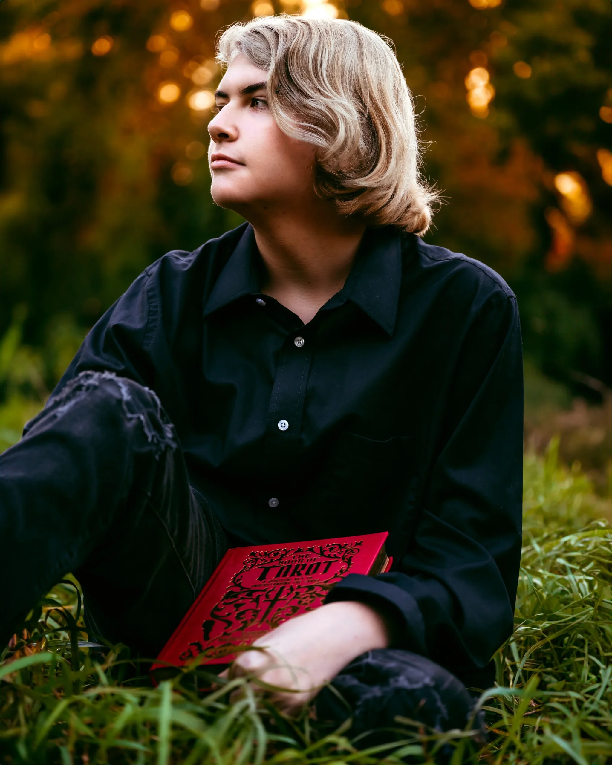 Person with medium-length blonde hair sitting in grassy area, wearing a black shirt and holding a red book.
