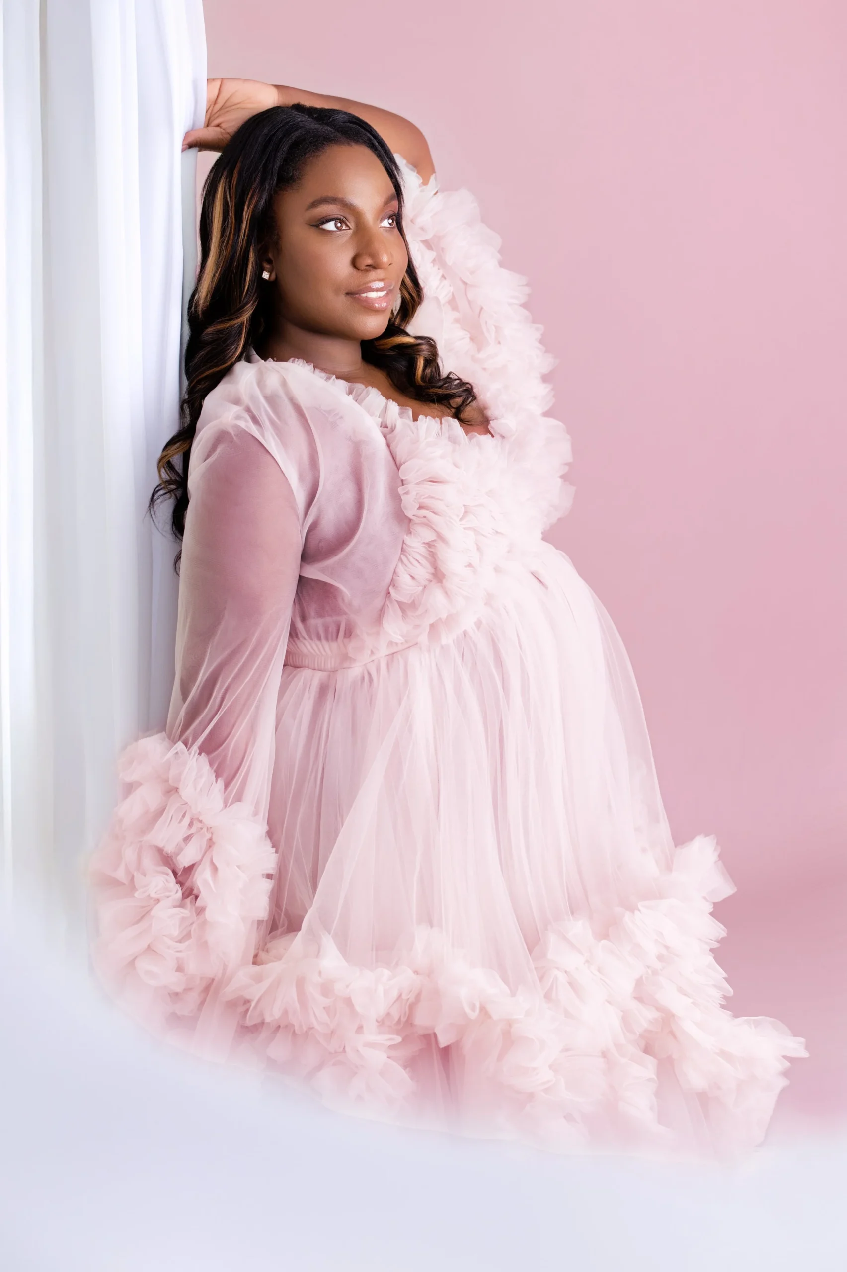A pregnant woman in a pink, ruffled, sheer dress sitting against a pink background.