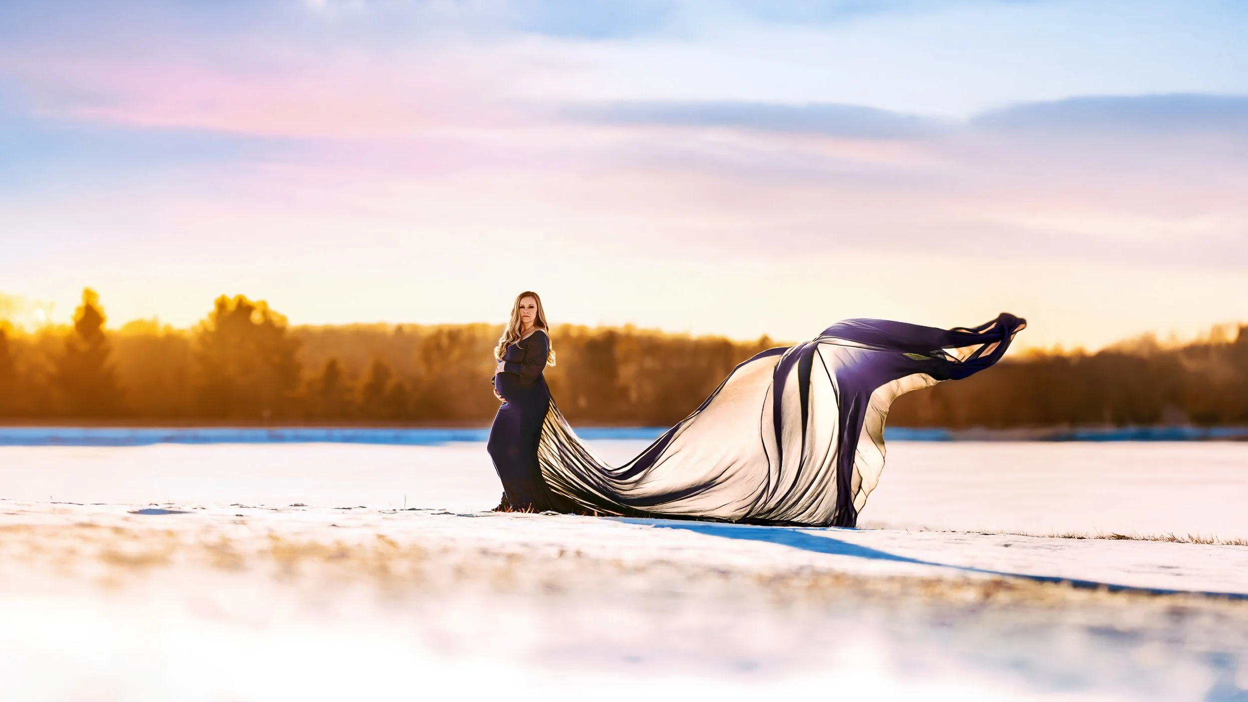 A pregnant woman standing on snow-covered ground at sunset, with a flowing black and beige dress. The background features trees and a colorful sky with pink and blue hues.