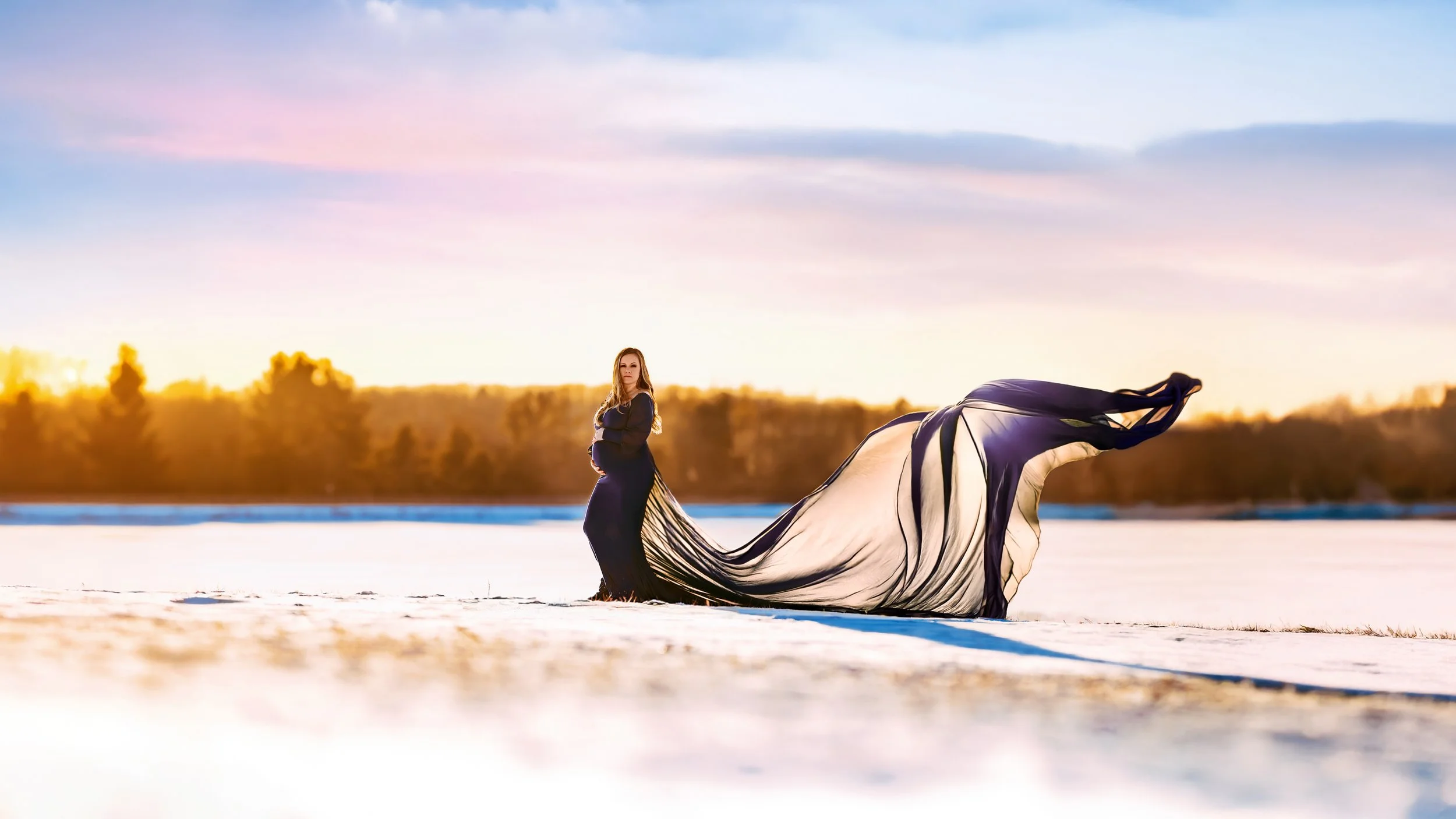 A pregnant woman in a long black dress posed on a snowy field at sunset, with a flowing black and beige cape billowing behind her.