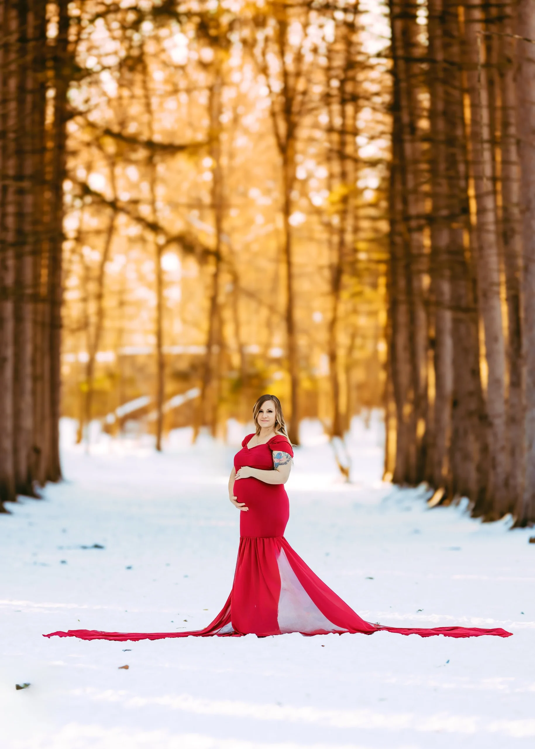 A pregnant woman in a red dress standing on snow in a forest with tall trees and warm golden sunlight.