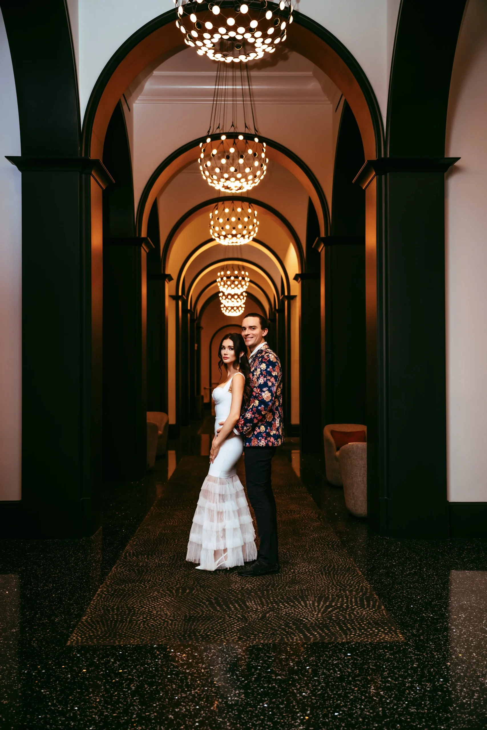 A couple standing in an elegant hallway with archways and chandeliers, the woman in a white dress and the man in a floral blazer.