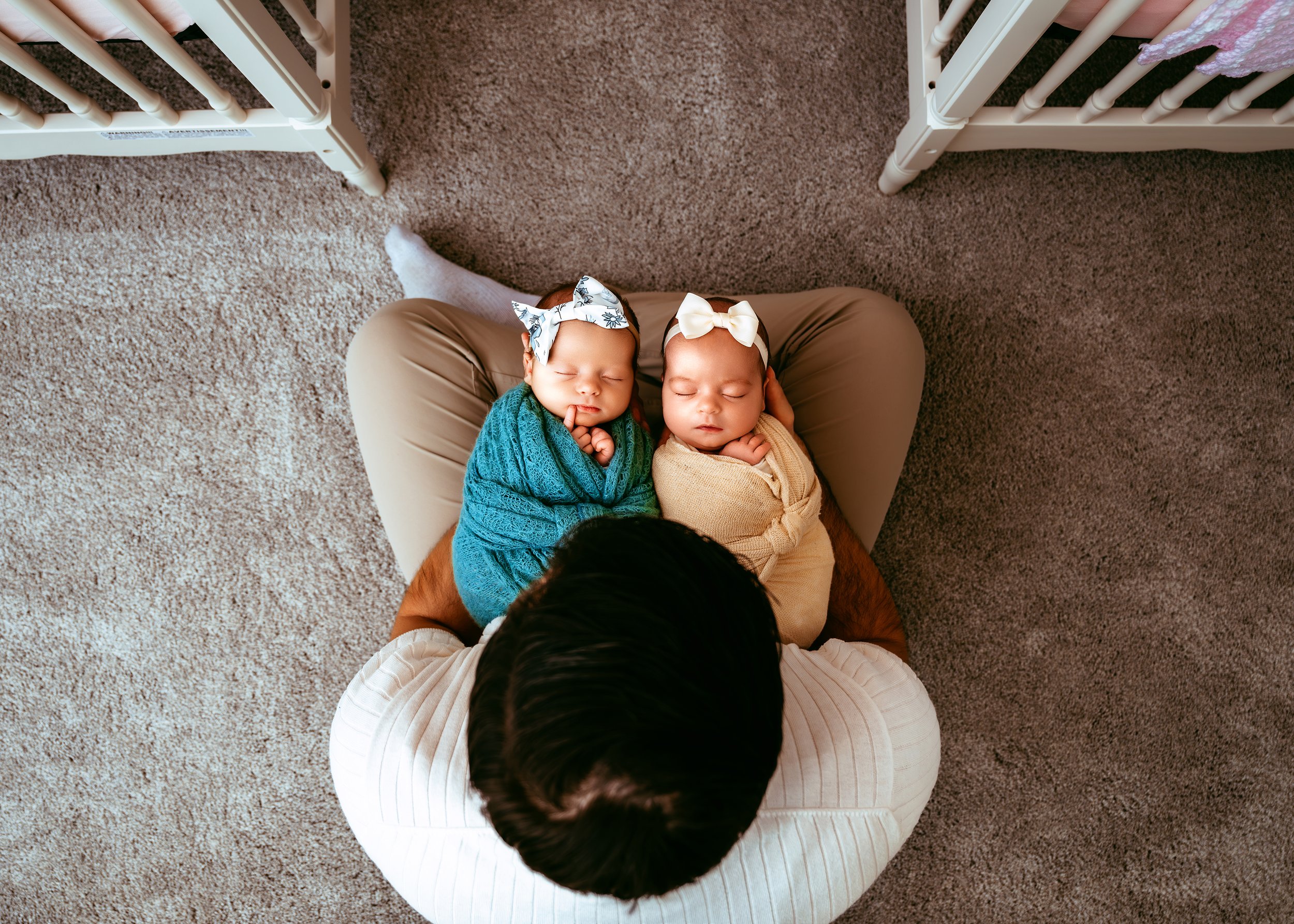 A person sitting on the floor with two swaddled newborns in their lap, each wearing a bow headband, next to two cribs.