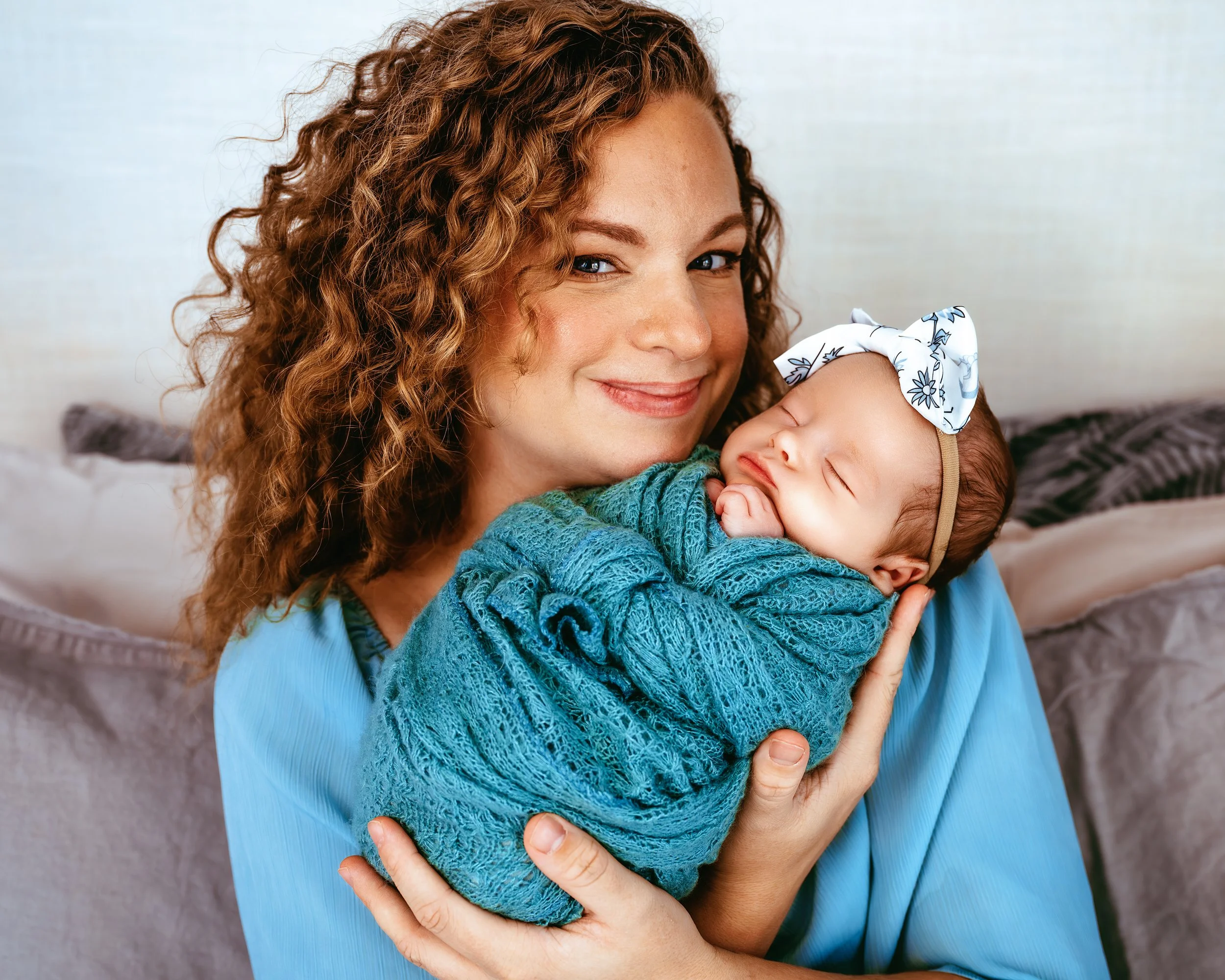A woman with curly hair smiling while holding a sleeping baby wrapped in a blue blanket.