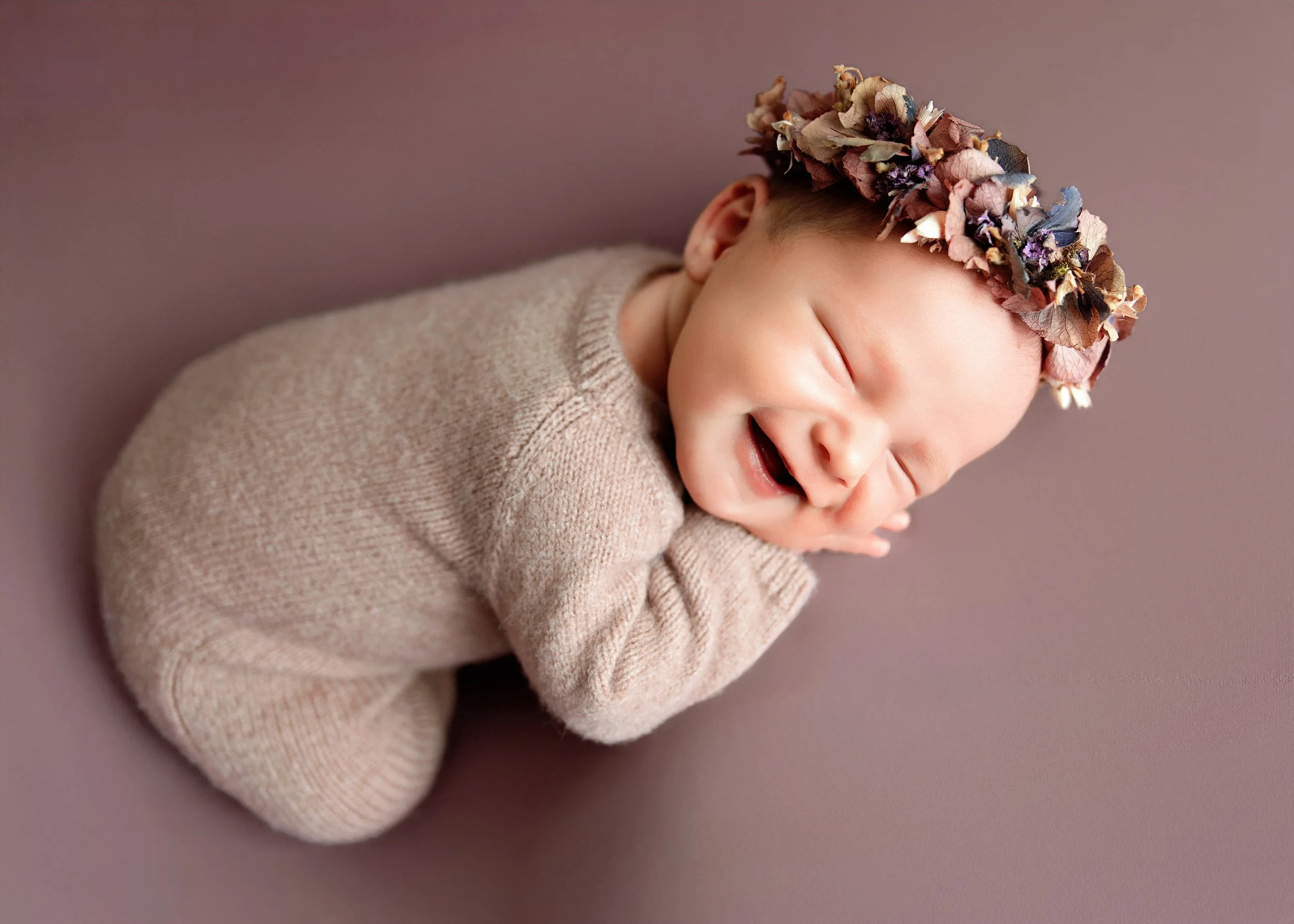 A smiling baby with a floral headband, wearing a beige sweater, lying on a mauve surface with eyes closed.
