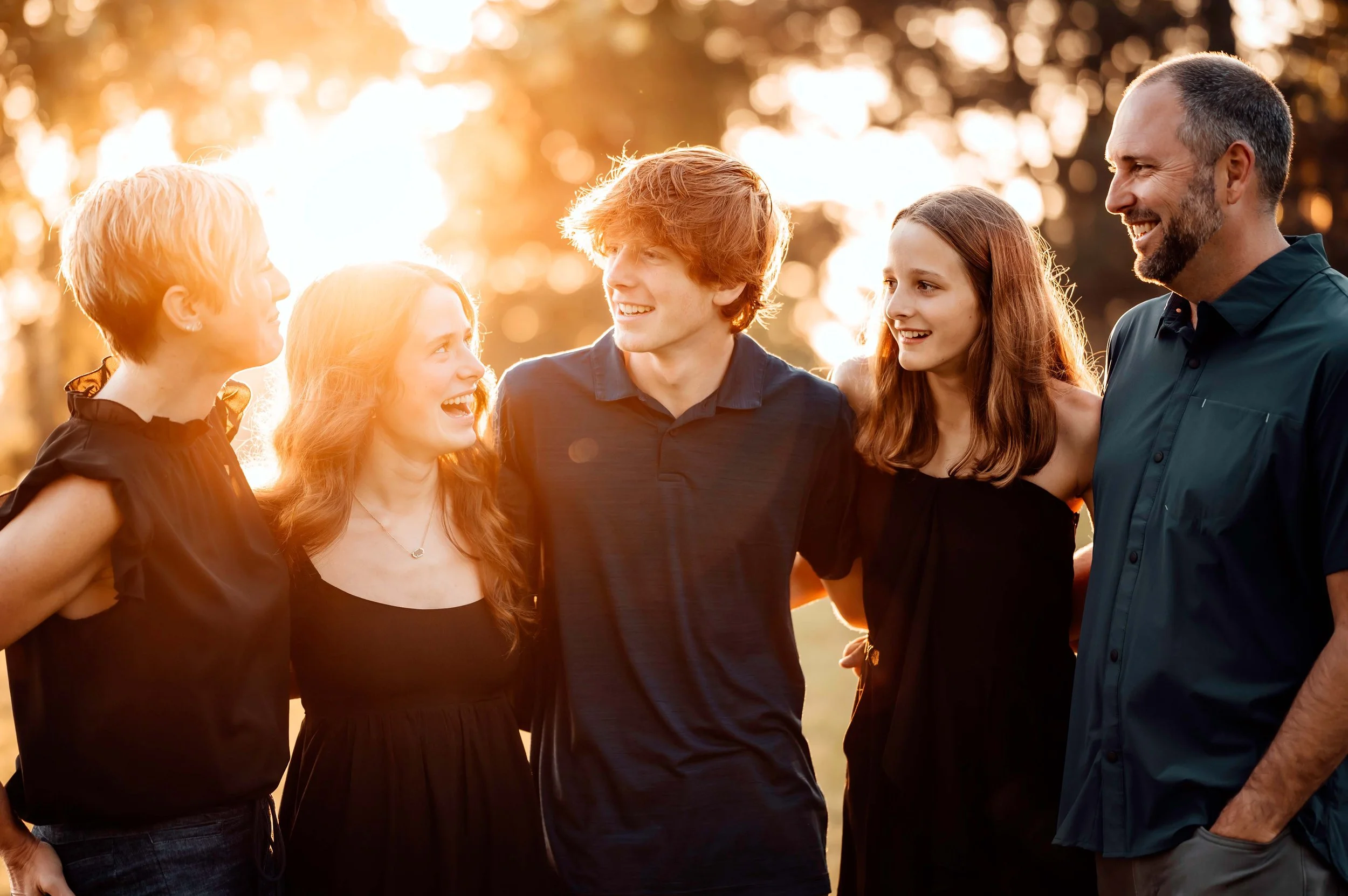 A group of five people standing together outdoors, smiling at each other in warm sunlight.