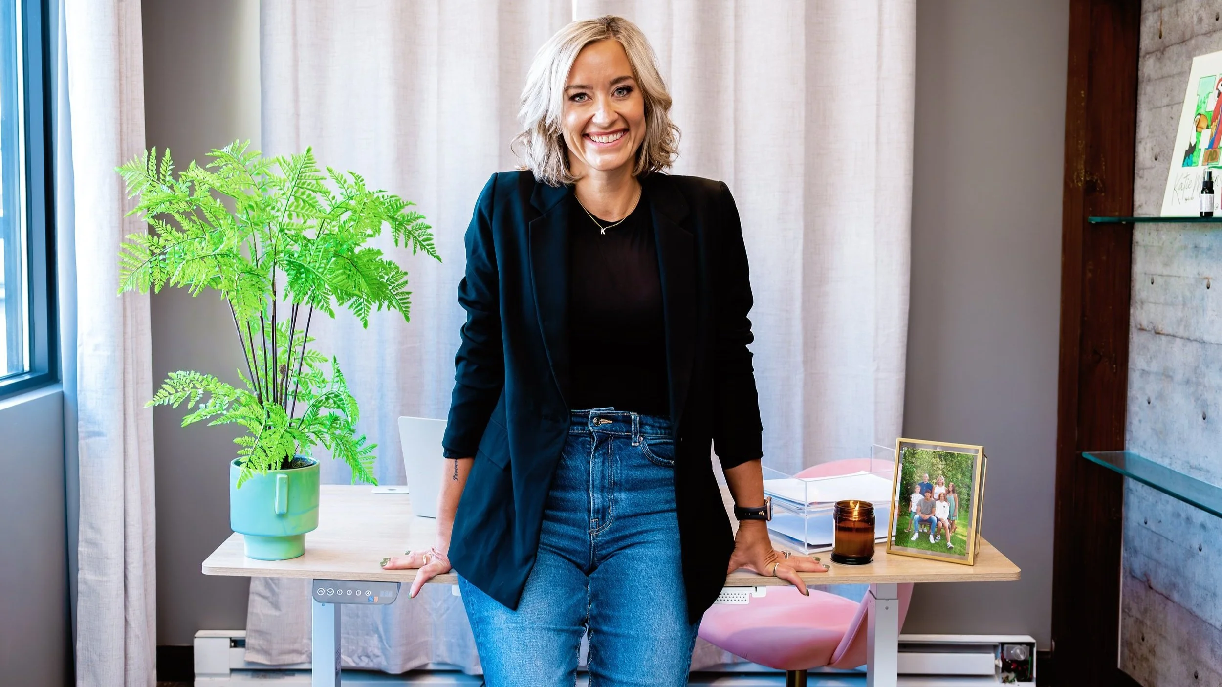 A smiling woman in a black blazer and jeans leans against a desk in a modern office space. A potted green plant and a framed family photo are on the desk, along with a lit candle. The background features curtains and a concrete wall with shelves.