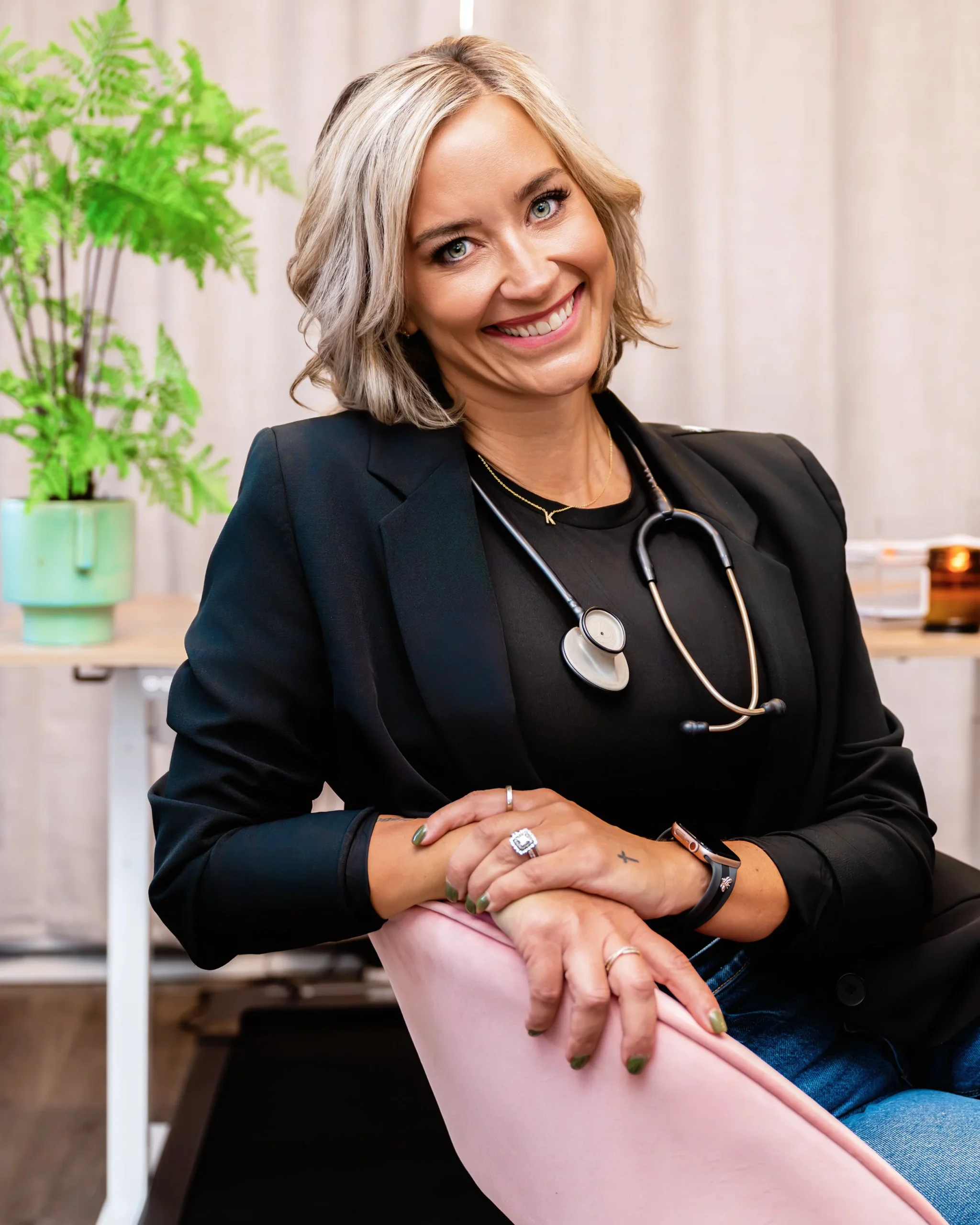 A smiling woman with blonde hair and a stethoscope around her neck, sitting in a pink chair in a clinical setting with a green plant in the background.
