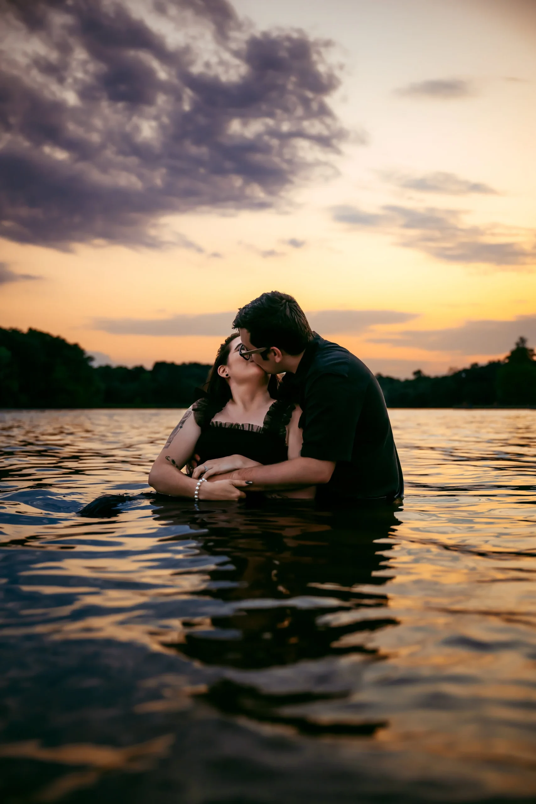 Couple kissing in a lake at sunset with orange and purple sky reflection on water.