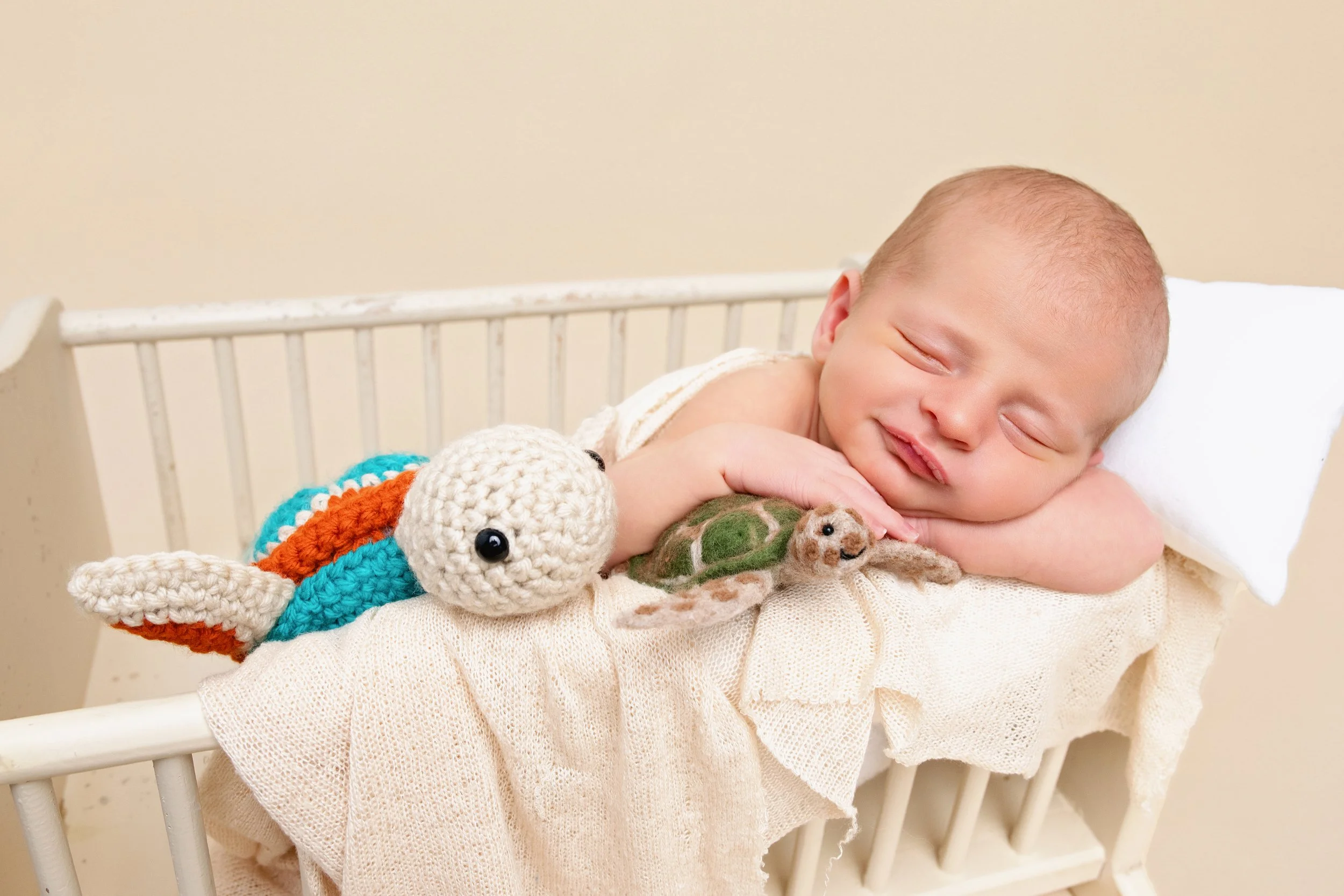 A smiling baby lying in a crib, resting on a pillow, with a crochet turtle and fish plush toys nearby.