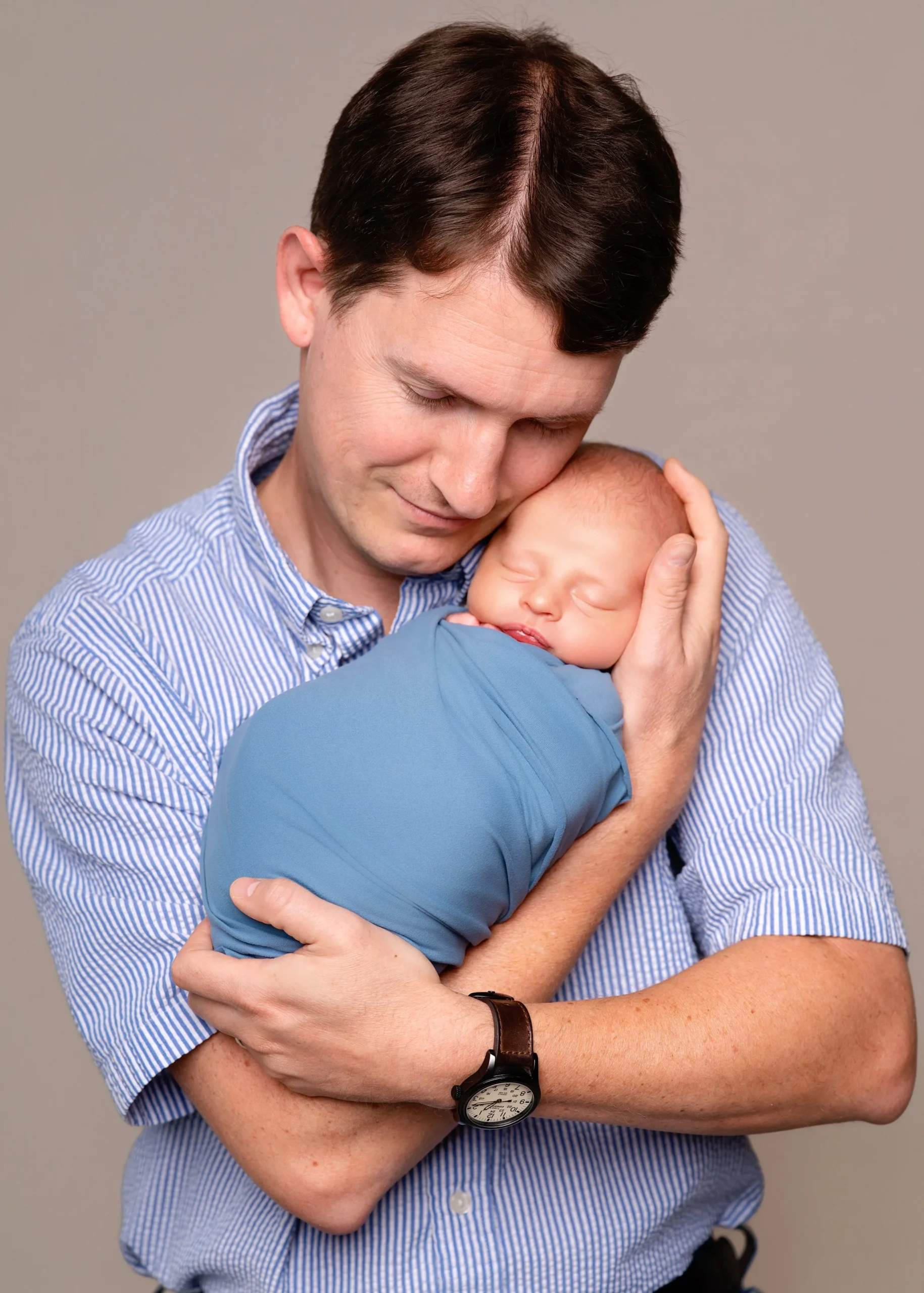 A man is holding a sleeping baby close to his chest with both arms, gently pressing his forehead against the baby's head in an affectionate embrace.