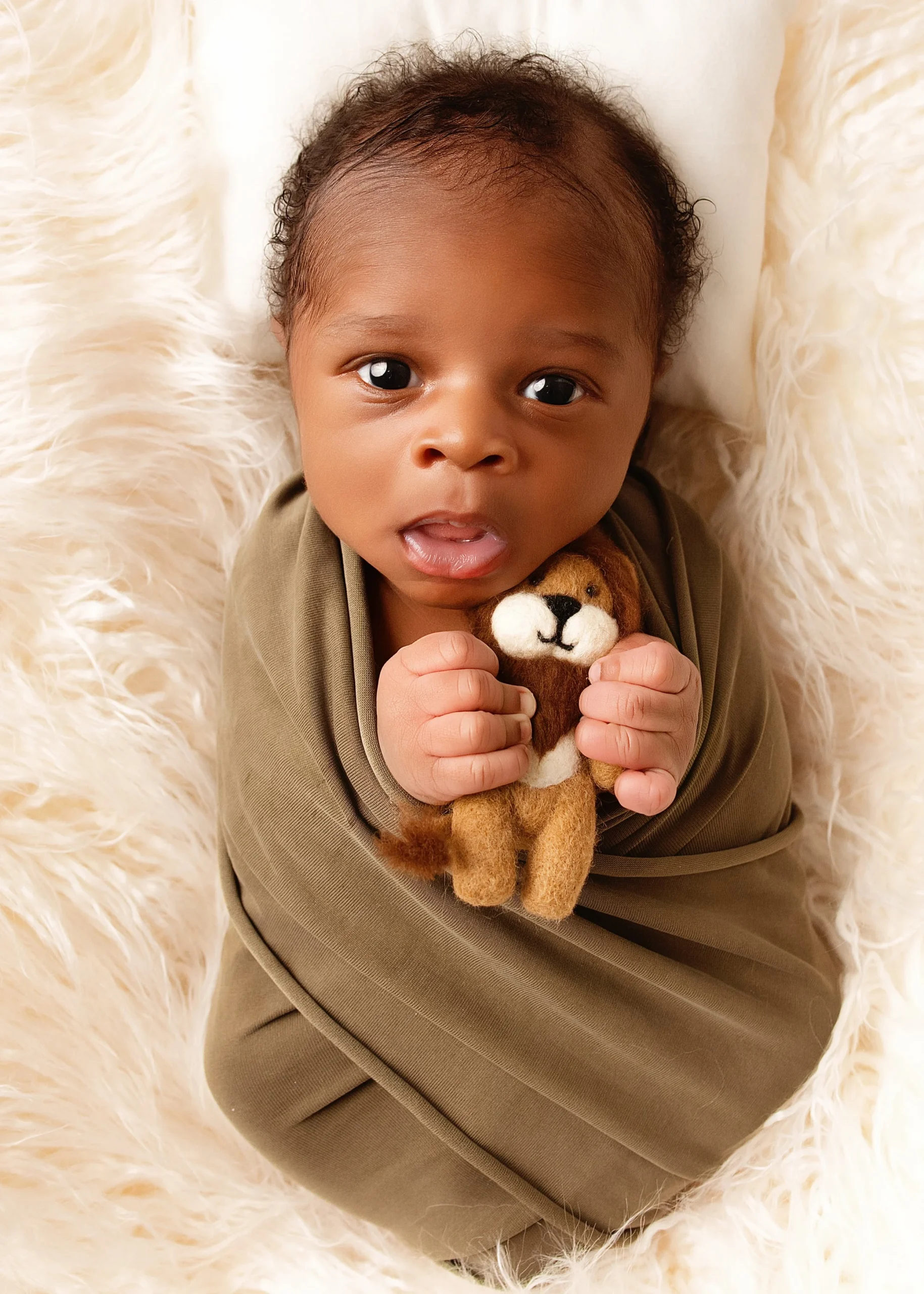 Young baby with dark hair lying on a fluffy white blanket, holding a small plush lion toy and looking up at the camera.