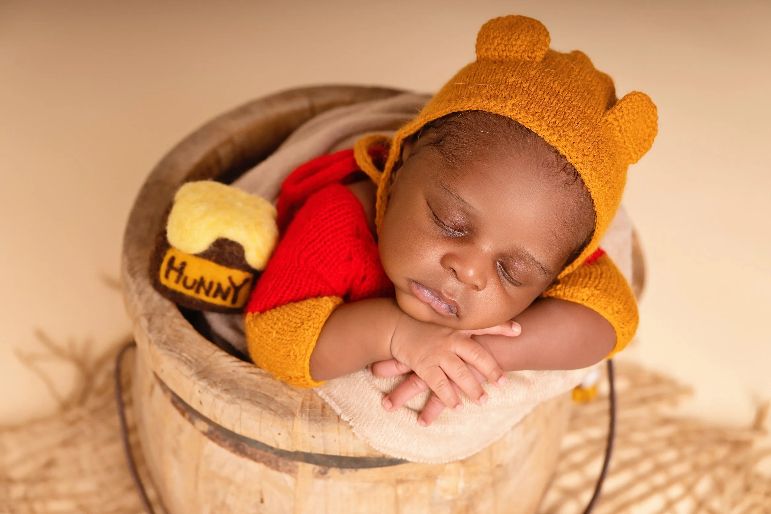 A sleeping baby with a yellow hat resembling a lion, resting on their arms inside a wooden bucket, dressed in red and yellow clothing, with a toy that says 'HUNNY' nearby.