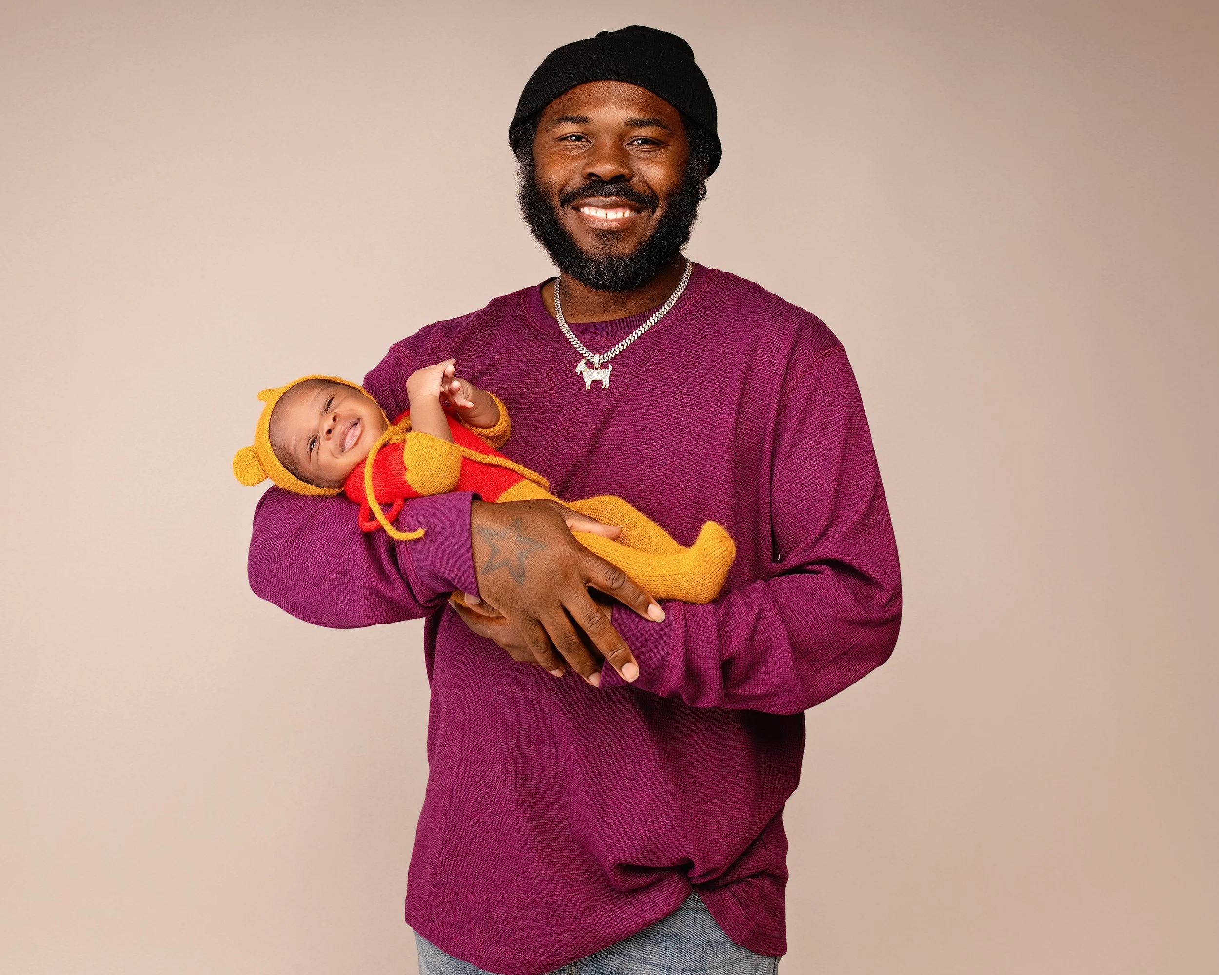 A man wearing a burgundy long sleeve shirt, black beanie, and a silver chain necklace with a dog pendant is smiling and holding a smiling baby dressed in a red and yellow animal costume with a bear hat.