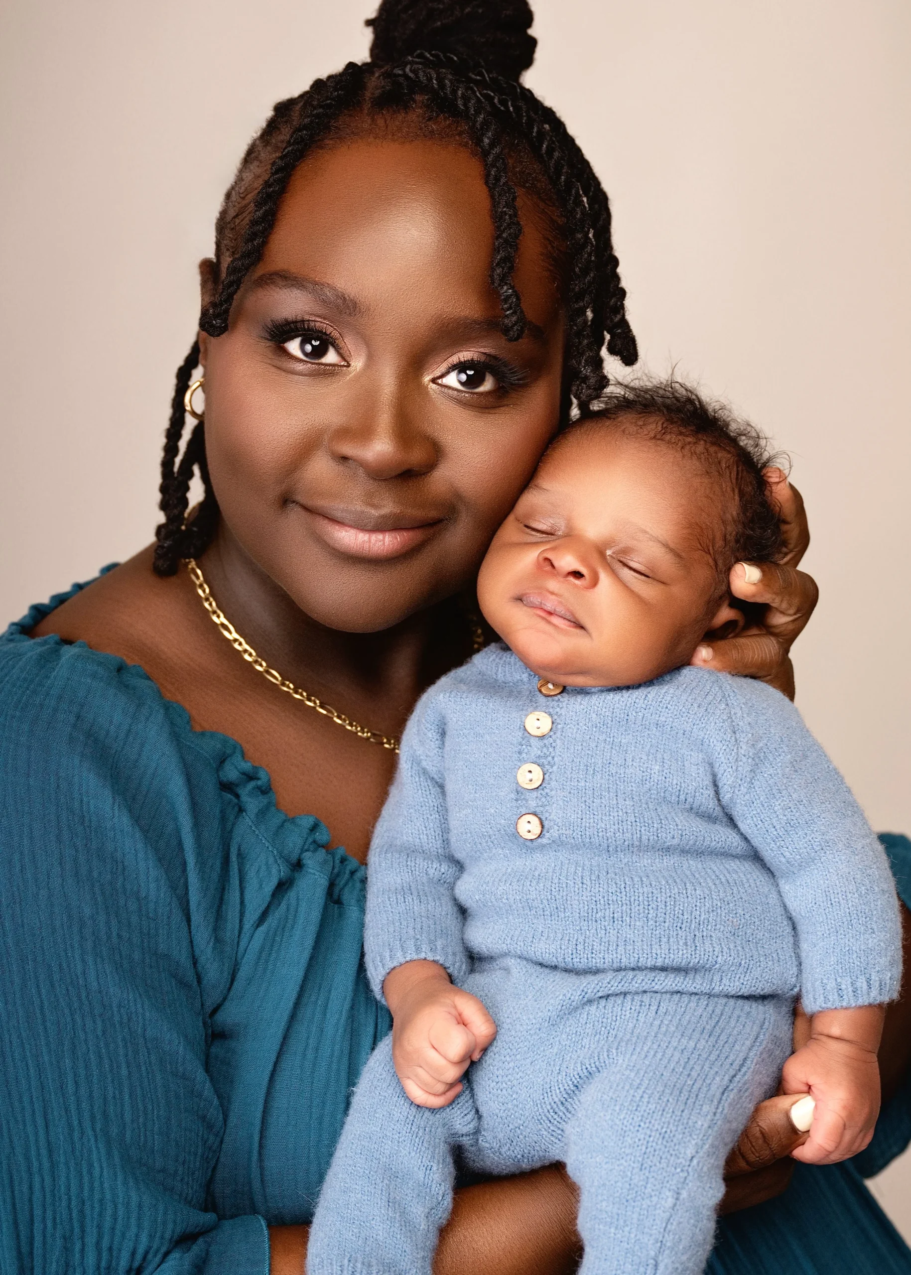 A smiling woman holding a sleeping baby dressed in a blue knitted outfit.