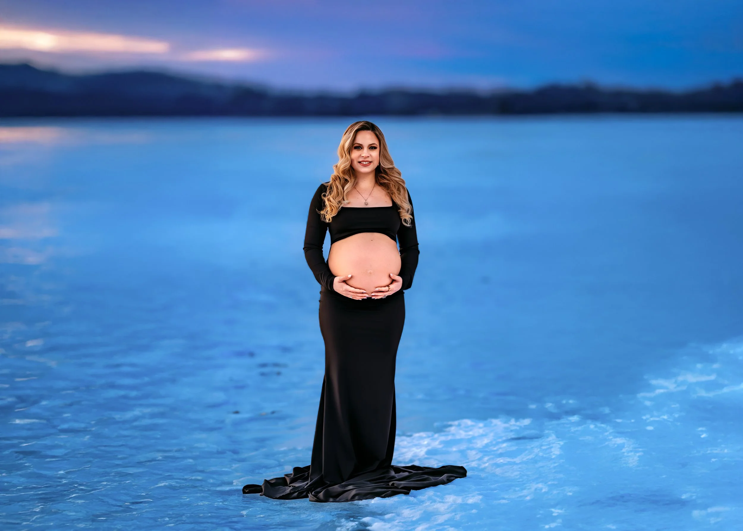 A pregnant woman with long blonde hair wearing a black dress standing in front of a body of water during sunset.