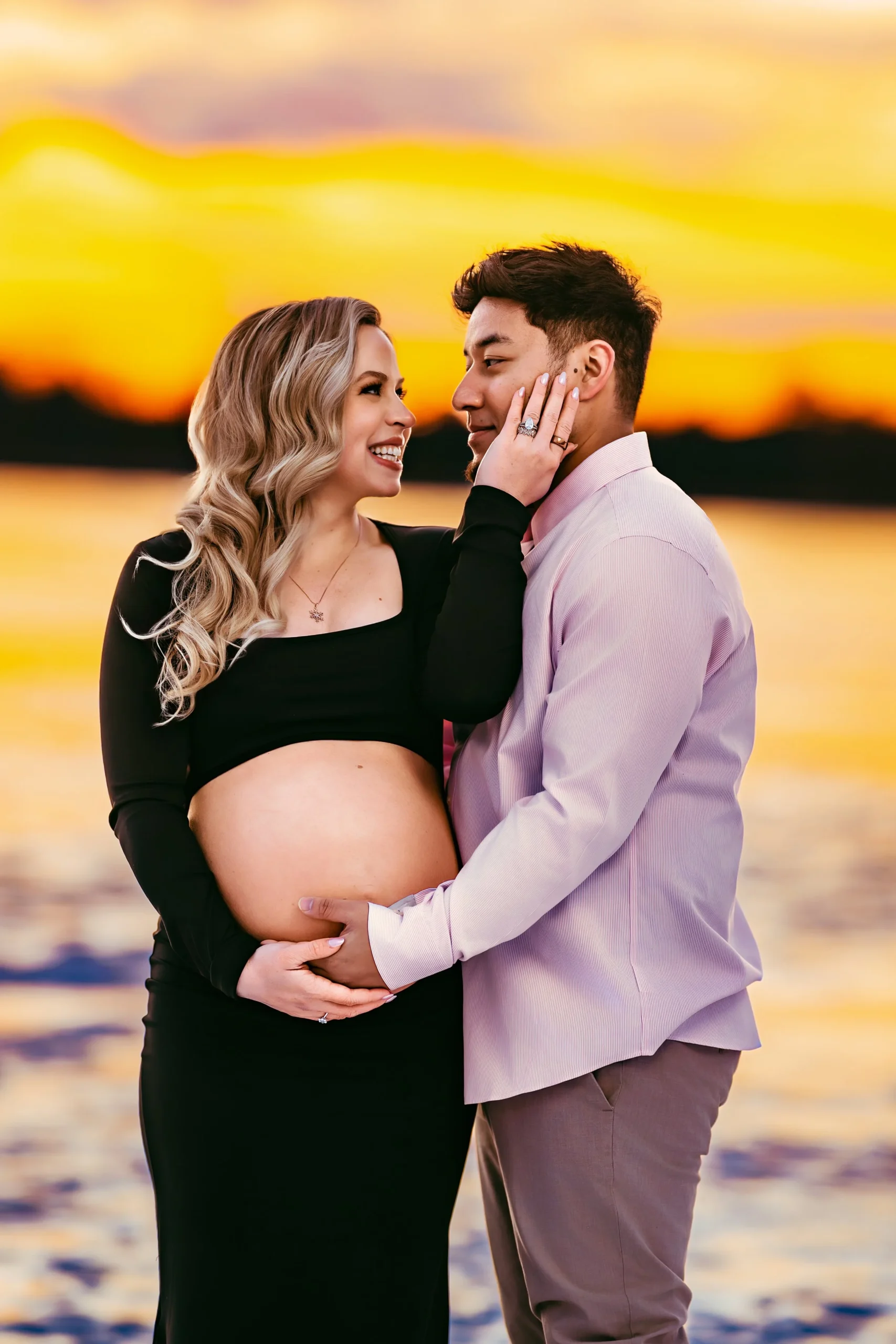 A pregnant woman and a man are standing close together at sunset by the water, with the woman touching the man's face and holding her belly, both smiling lovingly.